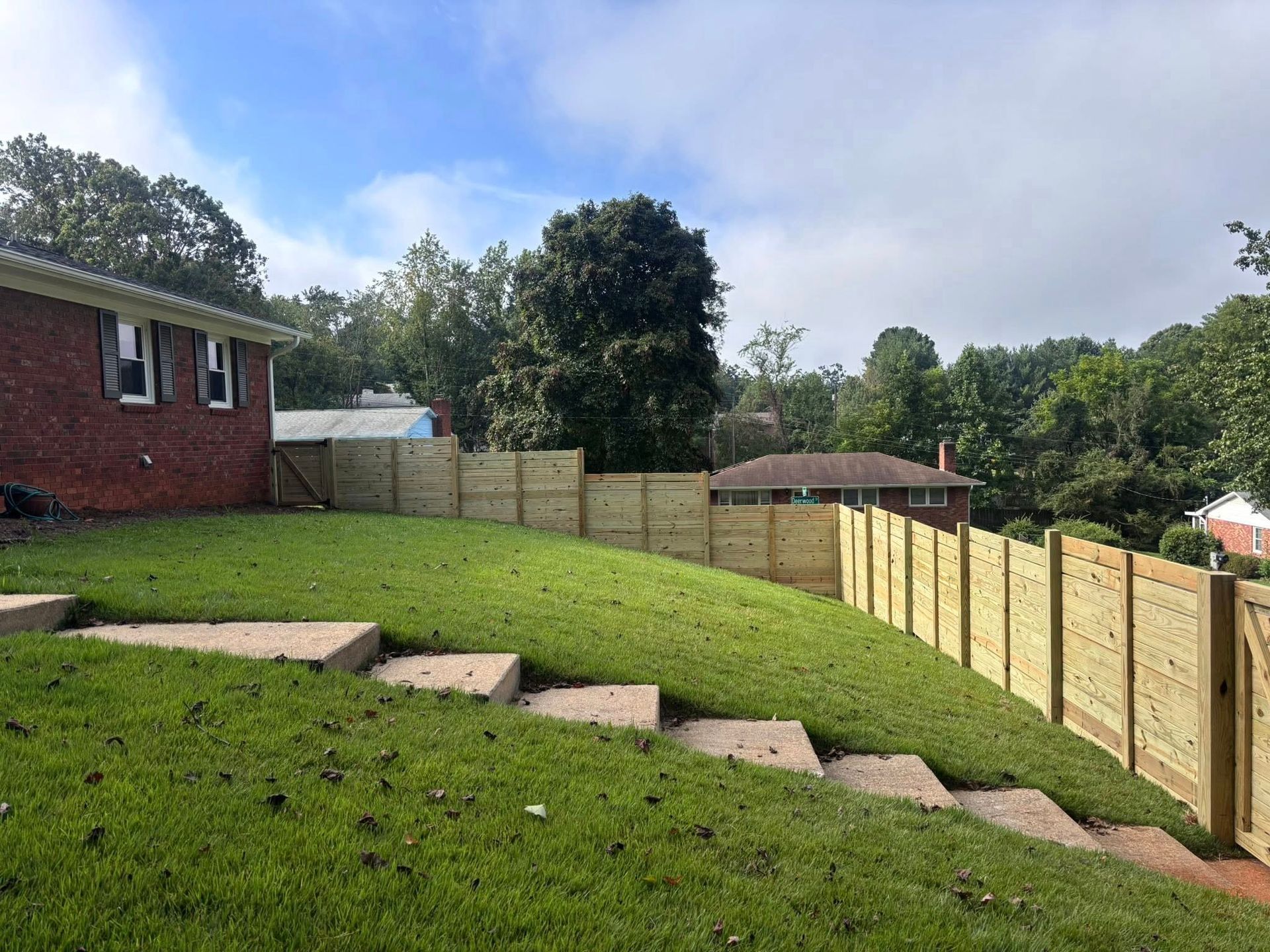 A newly installed wooden fence runs along a sloping grassy backyard next to a brick house with concrete stairs.