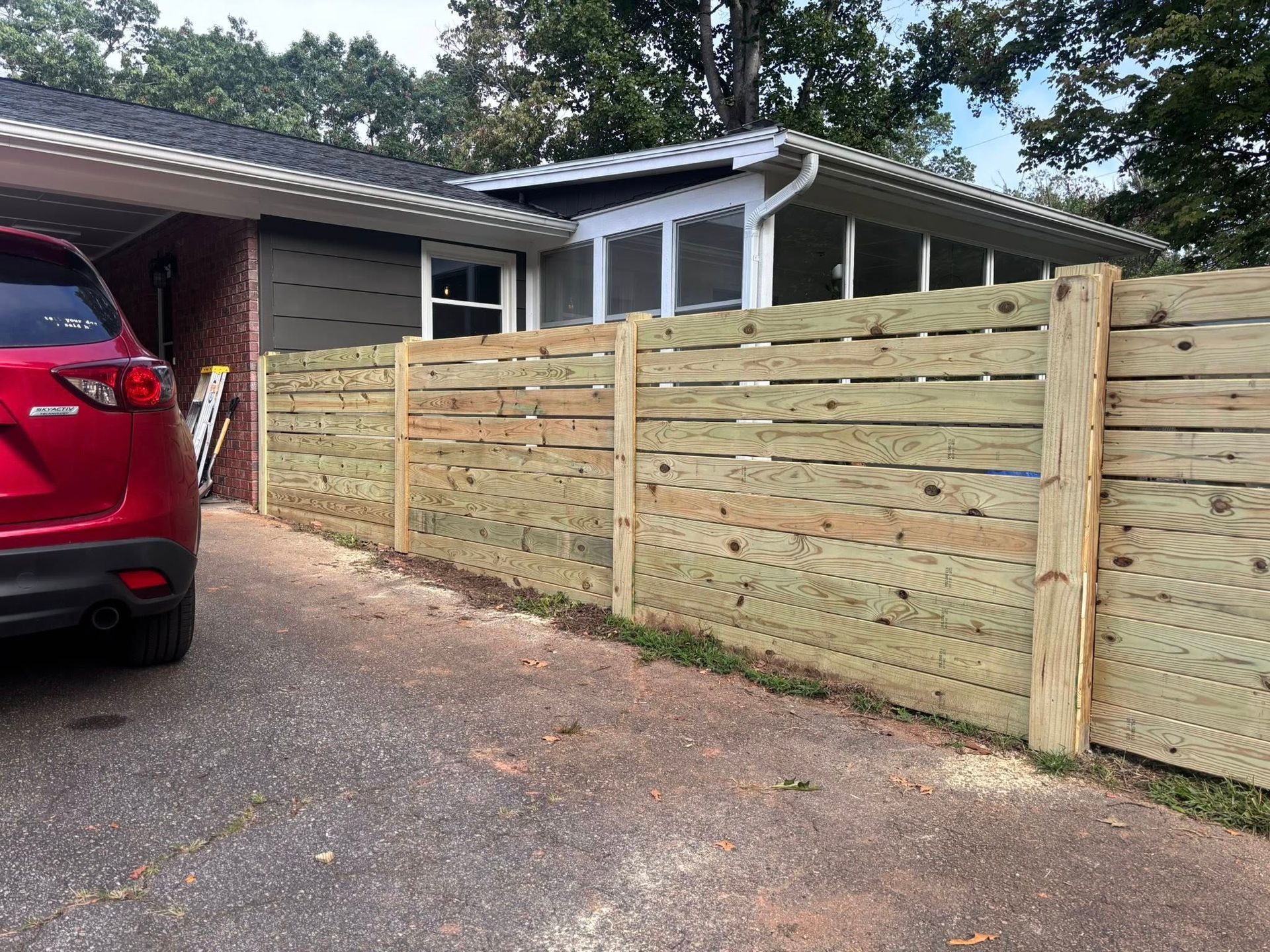 A newly installed horizontal wood slat fence runs alongside a driveway next to a house with a screened-in porch.