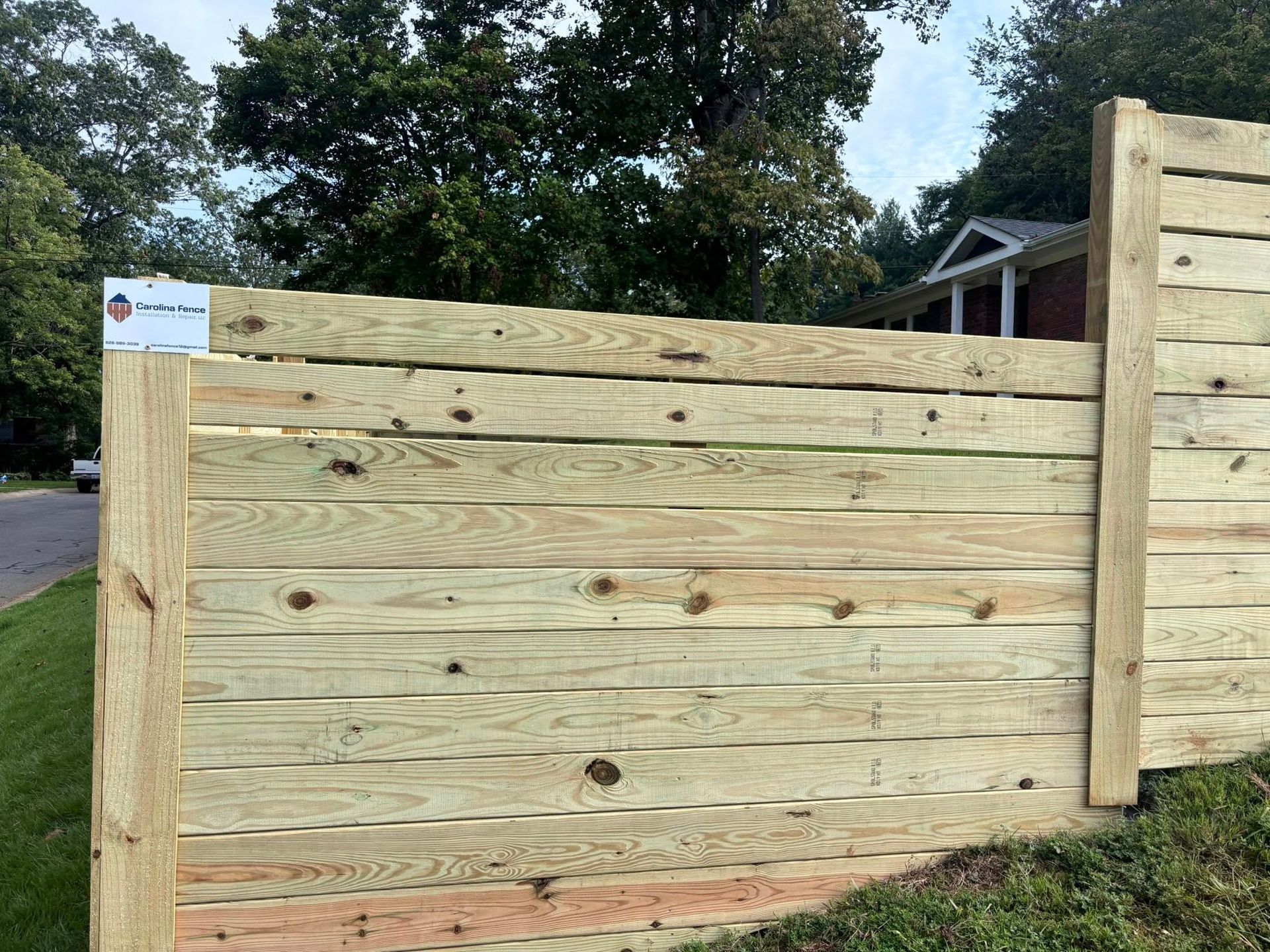 A wooden fence made of horizontal planks with vertical posts stands on a grassy slope with trees and a house in the back.
