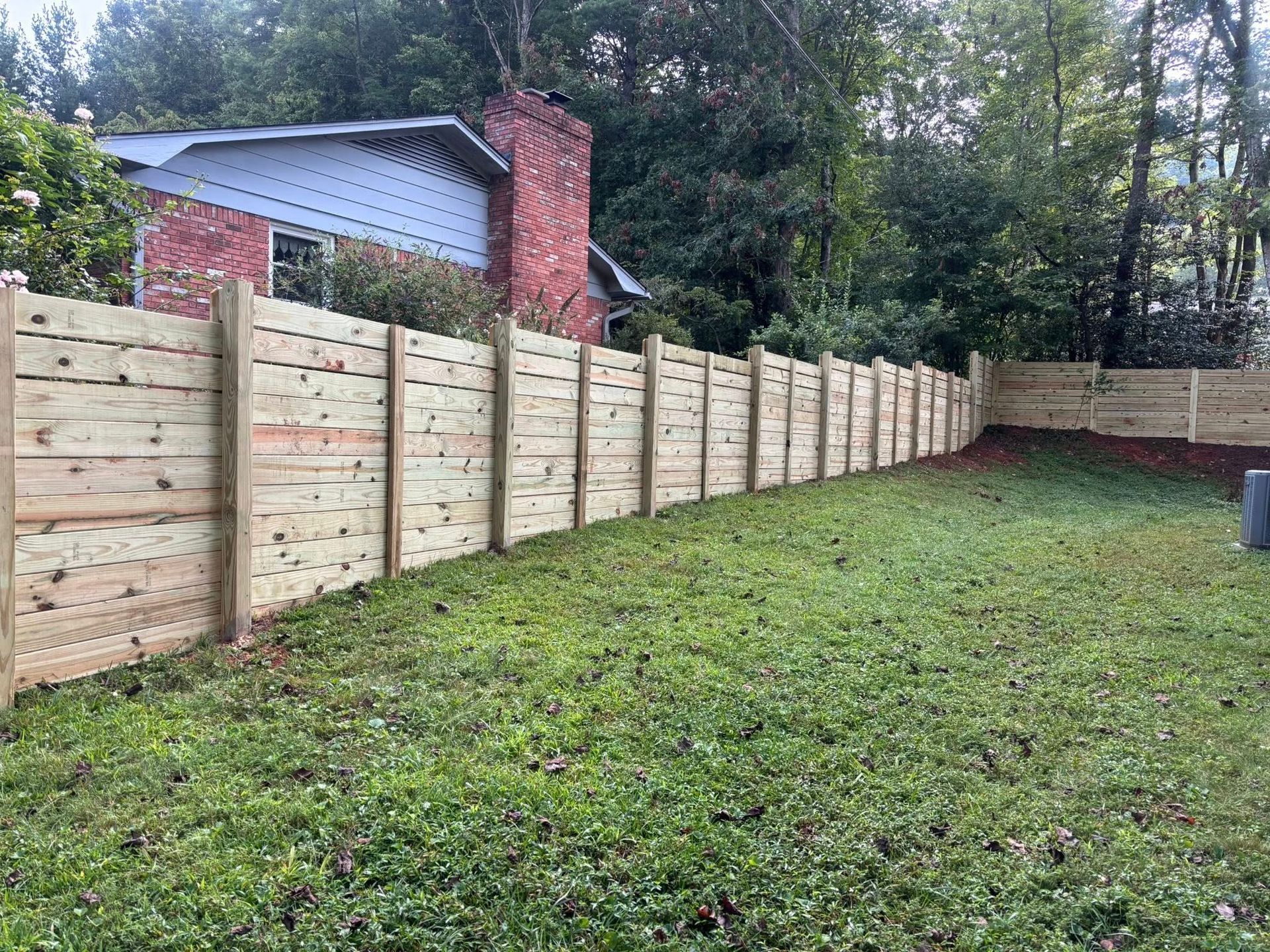 A horizontal wooden fence runs along the edge of a grassy yard, with a brick and light-blue house in the background.