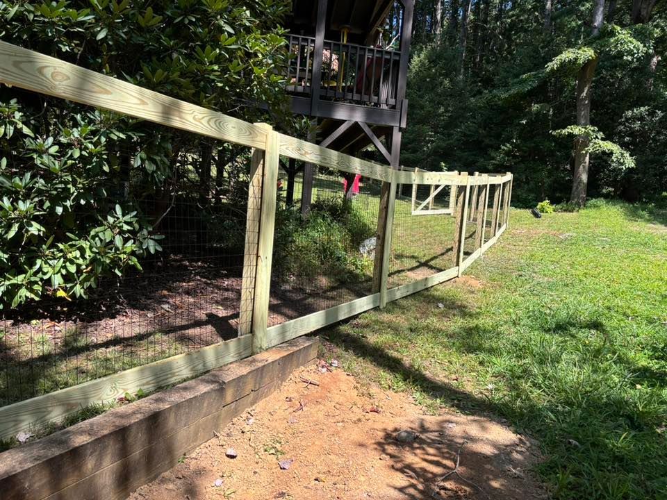 A new wooden post-and-rail fence stands along a stone retaining wall in a grassy, wooded yard.