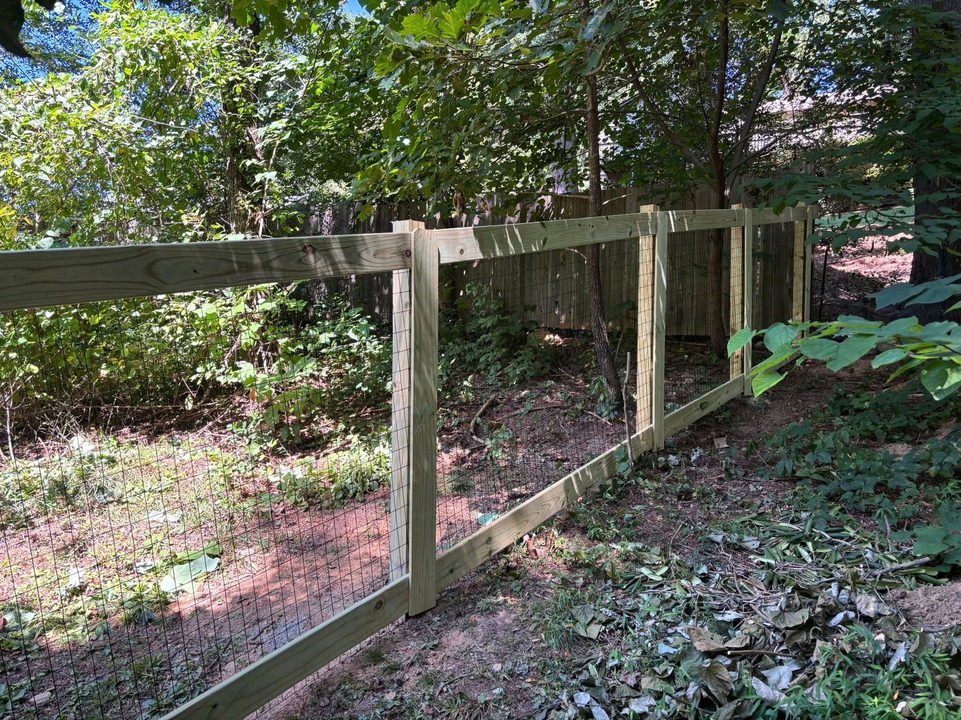 A newly installed wooden fence frame with horizontal rails and vertical posts stands in a wooded area with dense foliage.