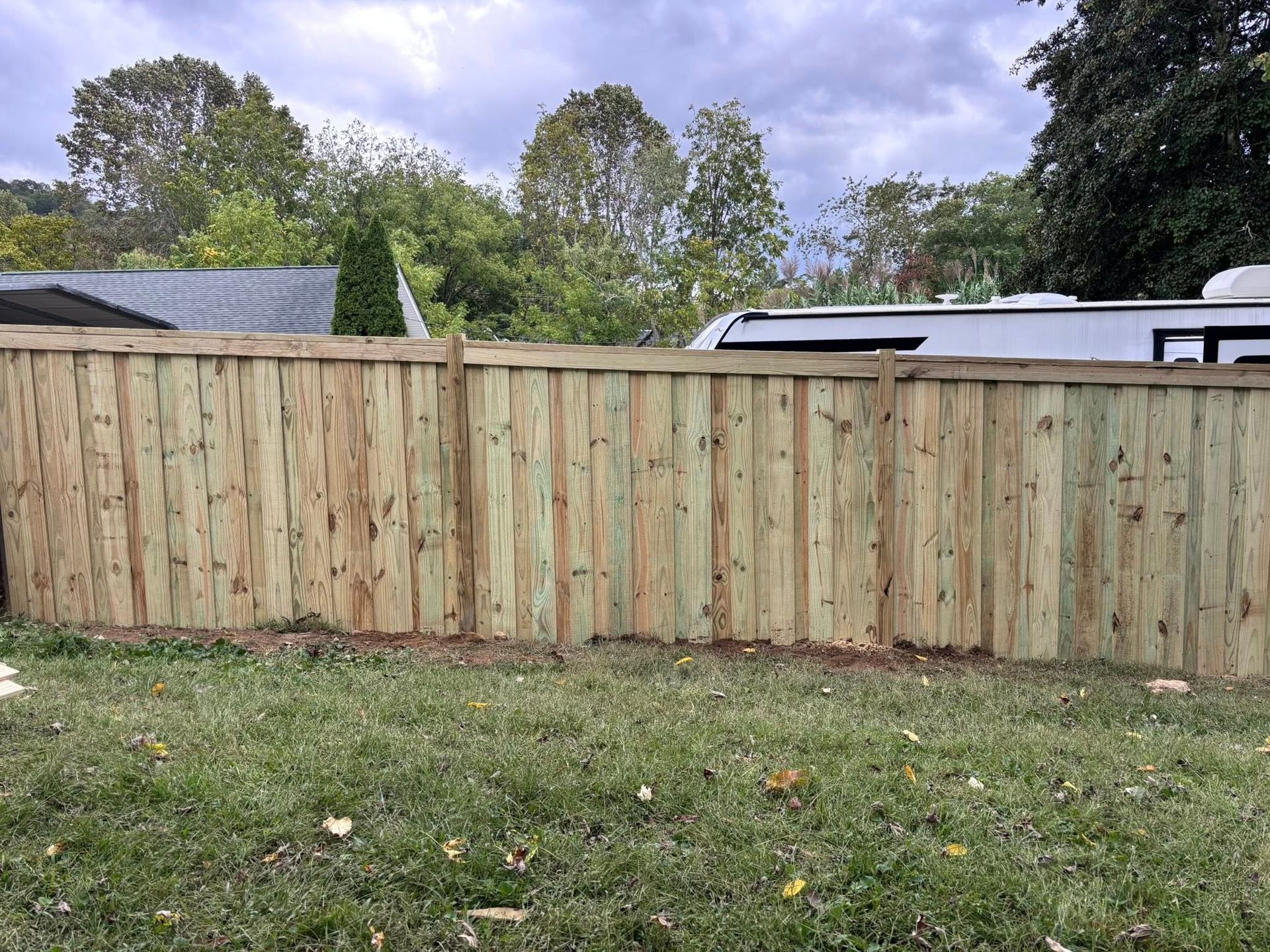 A tall wooden privacy fence made of vertical slats stands in a grassy yard under a cloudy sky.