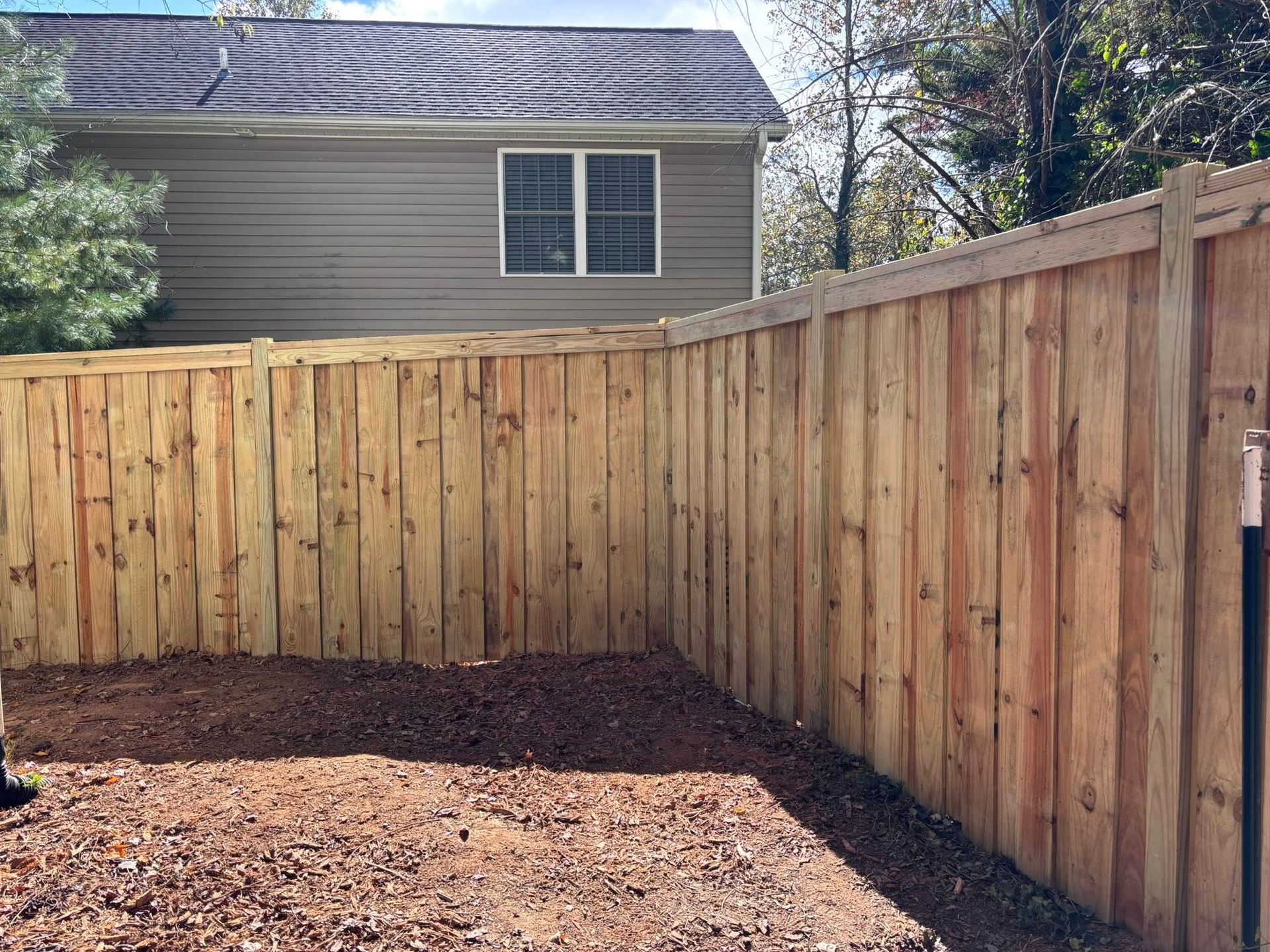 A wooden privacy fence encloses a dirt backyard in front of a gray, two-story house with two windows.
