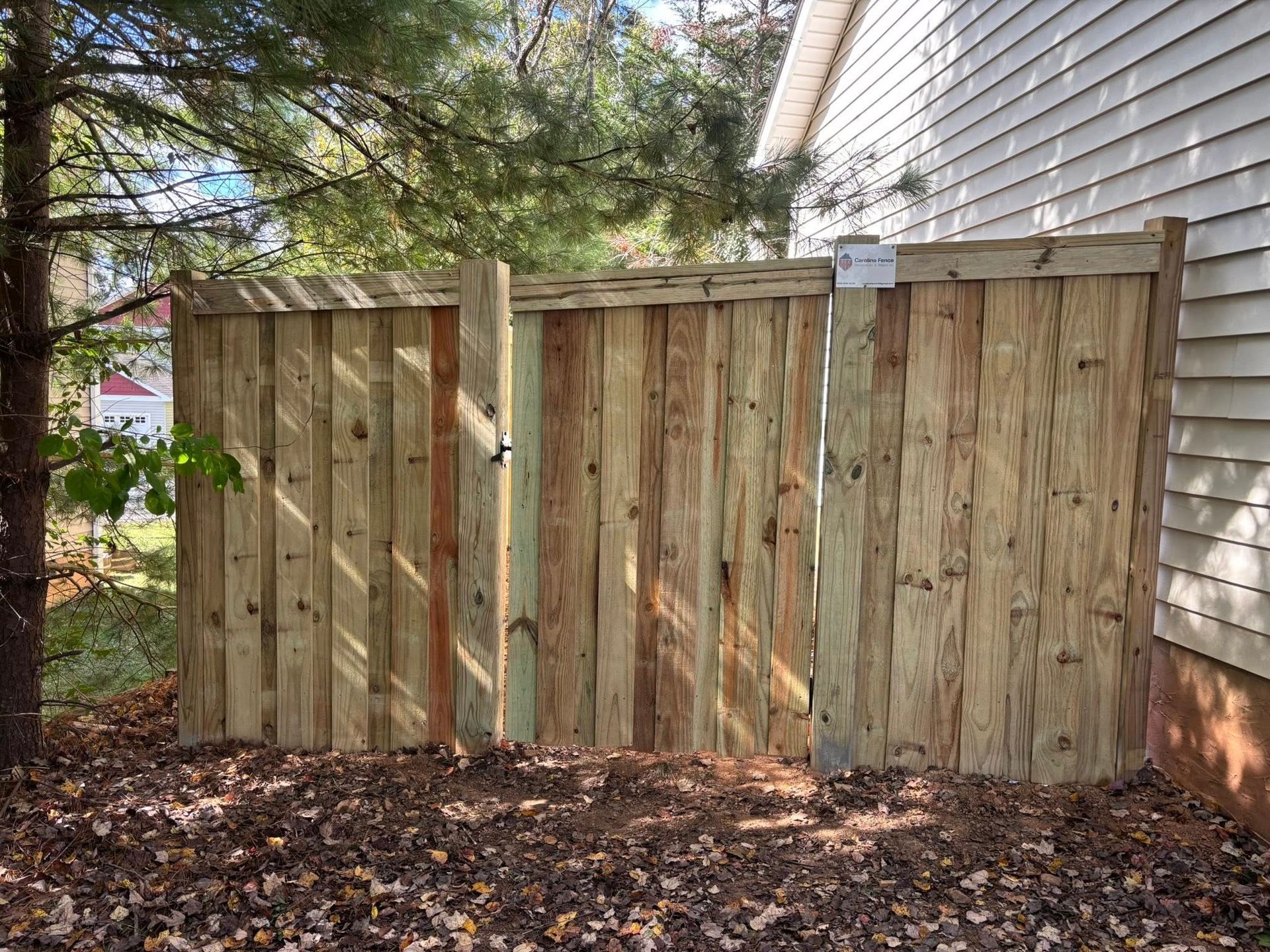 A new wooden privacy fence installed next to the side of a house, set against a background of trees and fallen leaves.