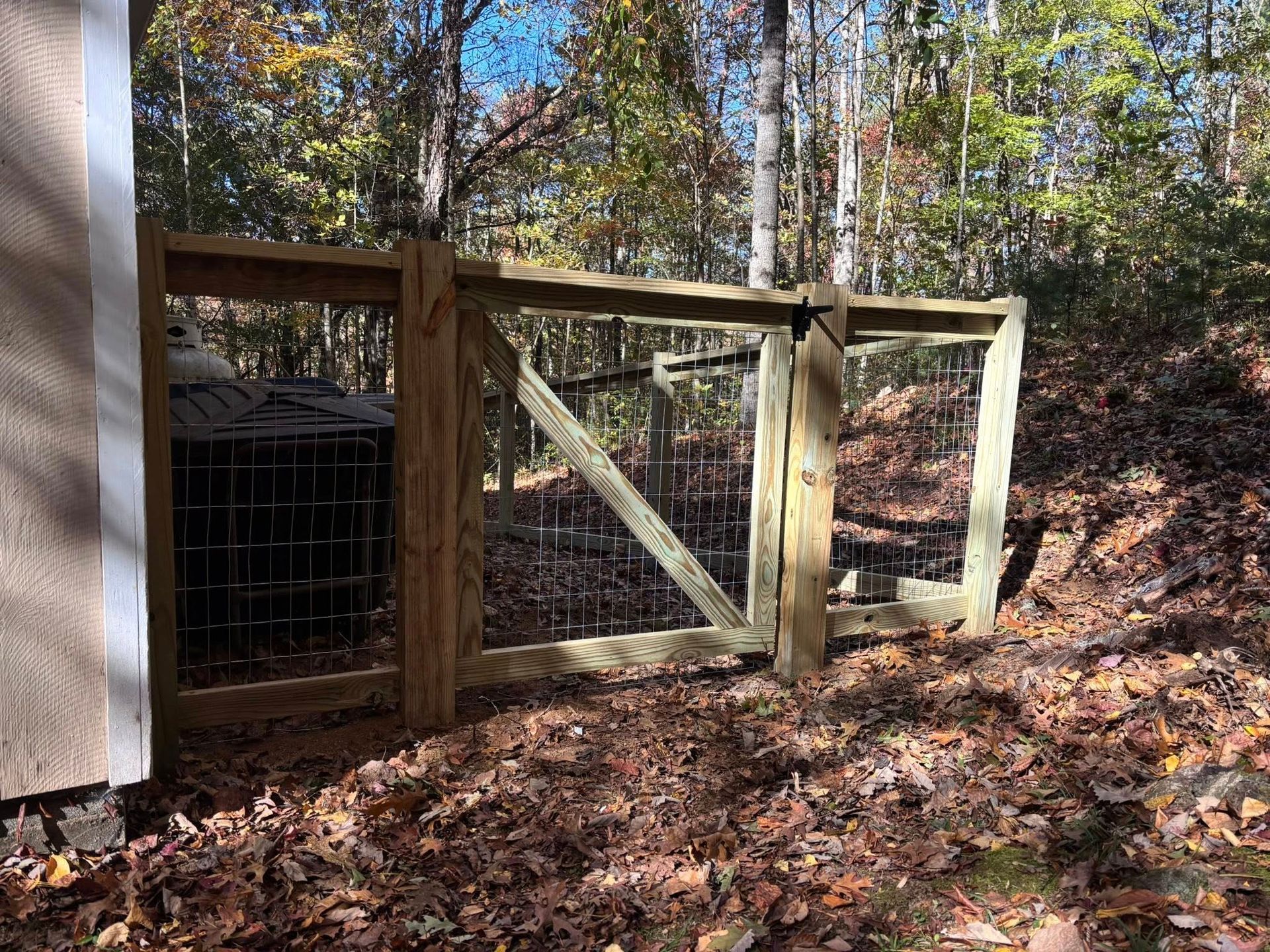 A newly constructed wooden fence with metal wire mesh, installed along a wooded slope next to a shed.