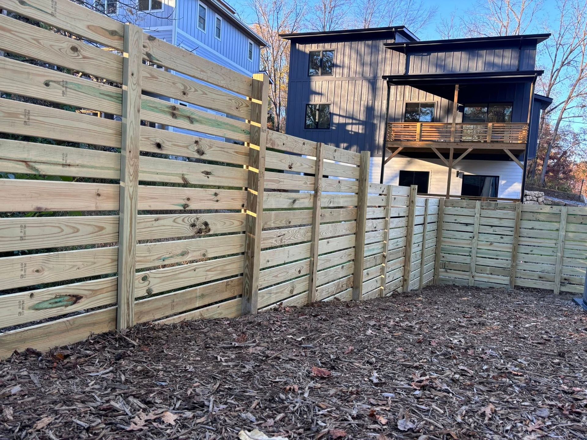 A tall, horizontal wooden privacy fence runs across a backyard covered in dry brown leaves near a multi-story house.