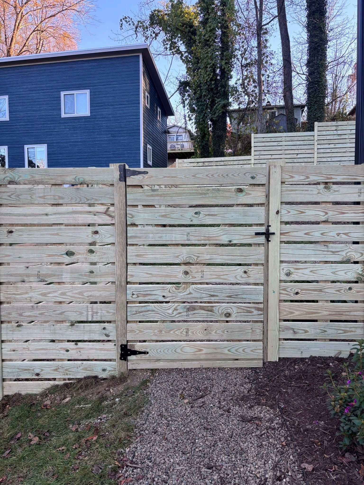 A wooden gate centered in a horizontal slat fence leads to a gravel path with a blue house visible in the background.