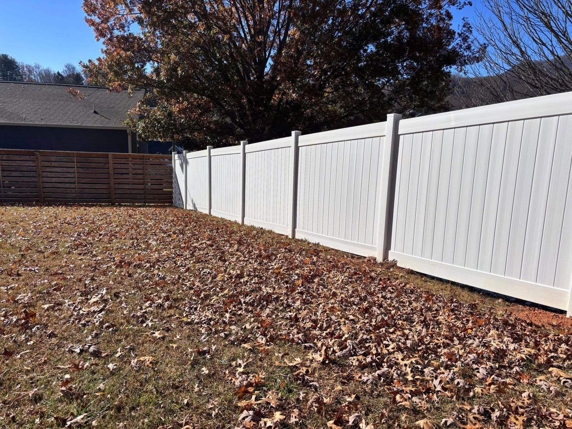 A white vinyl privacy fence lines a backyard covered in fallen autumn leaves under a clear blue sky.