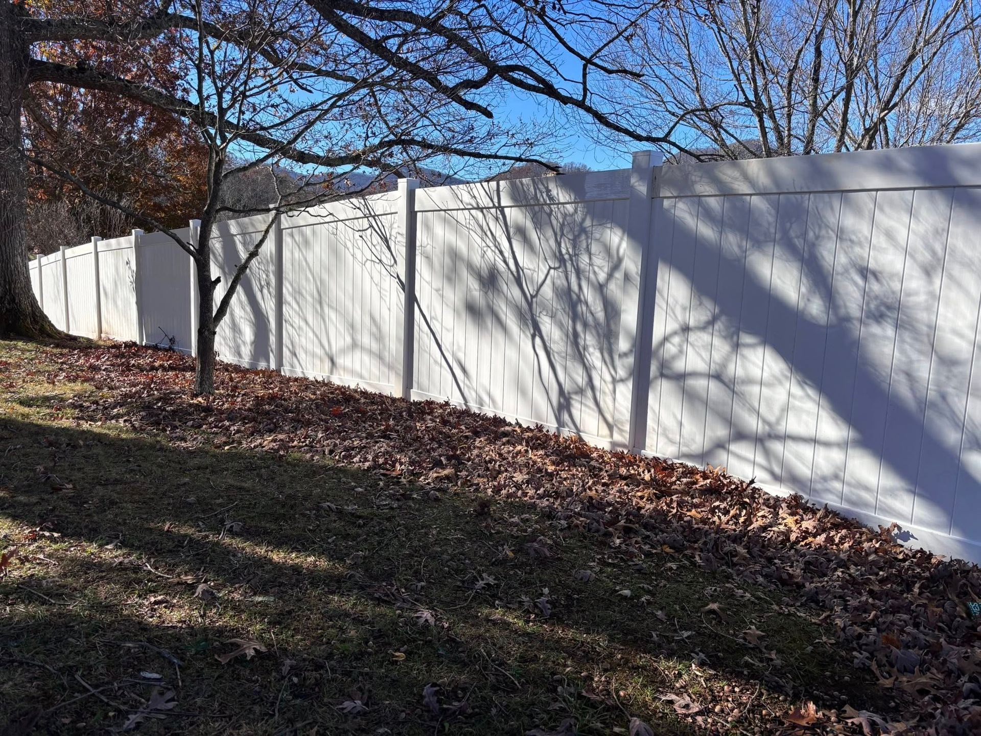 A white vinyl privacy fence runs along a grassy slope covered with autumn leaves under a clear blue sky.