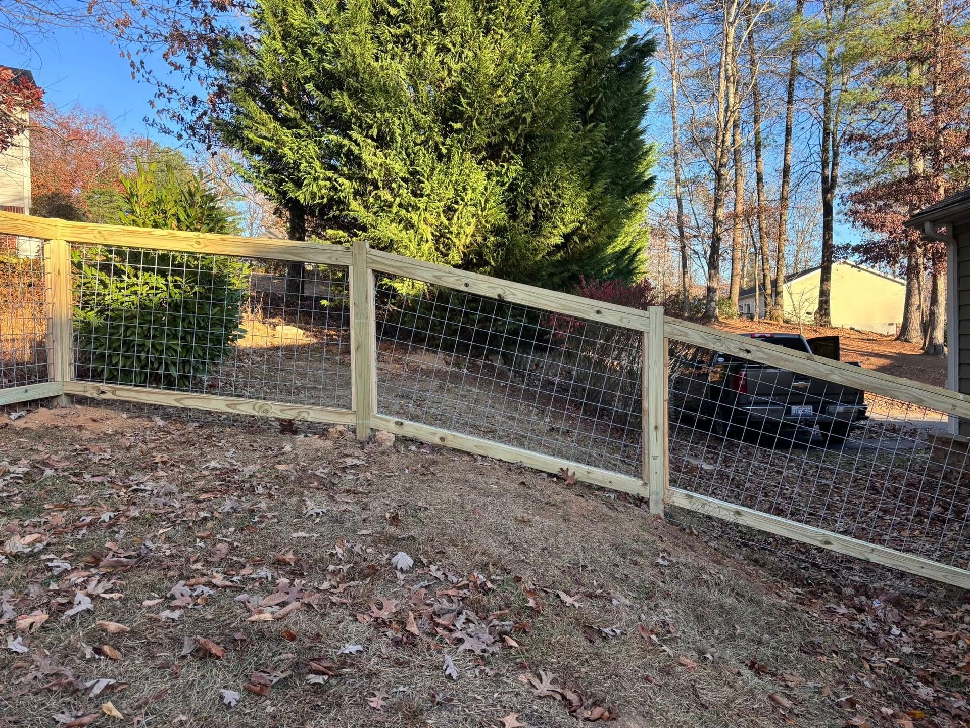A newly installed wooden post-and-rail fence with wire mesh in a grassy yard, with trees and a parked vehicle in the back.