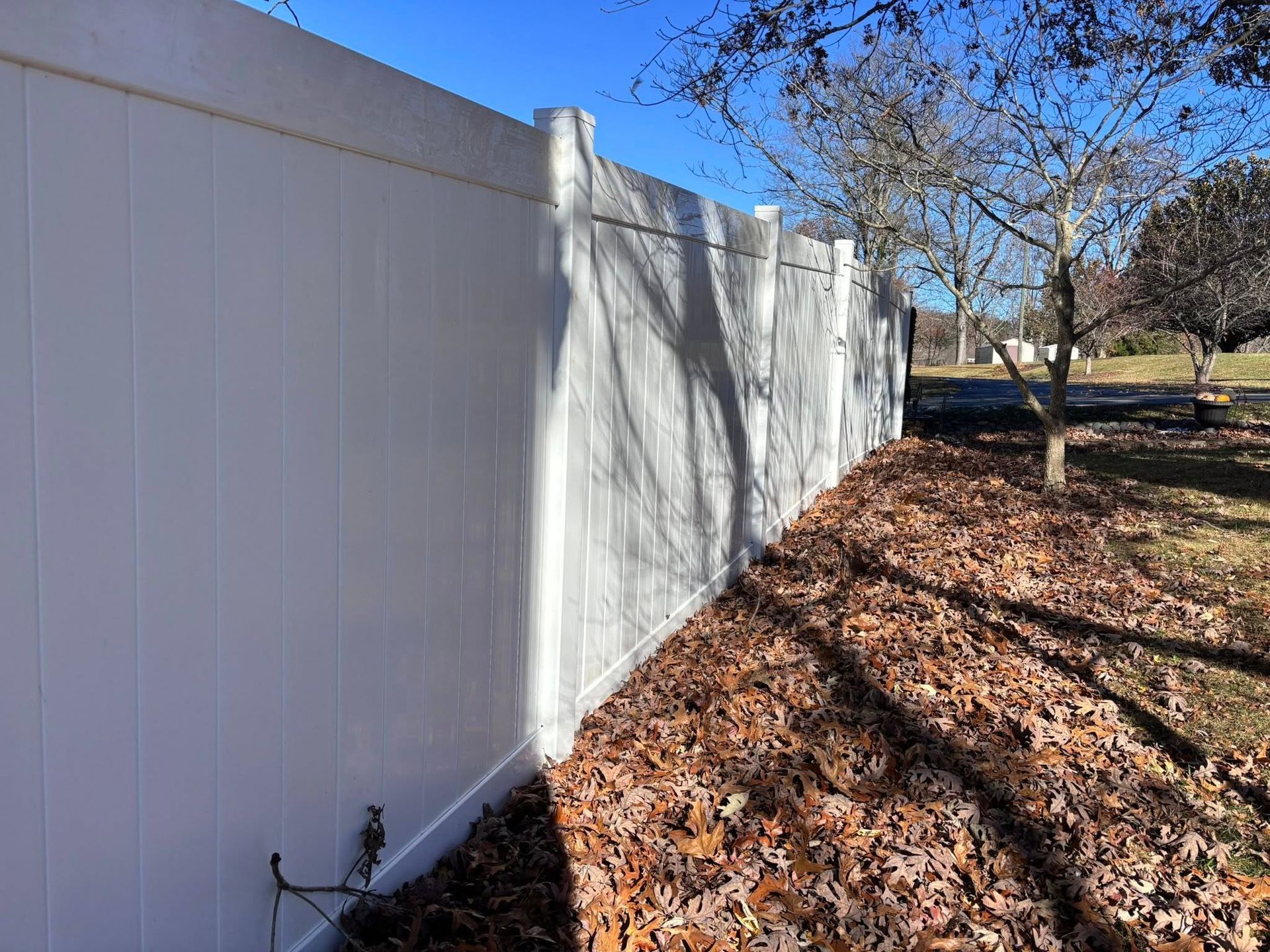 A tall white vinyl privacy fence stands outdoors next to a ground covered in brown autumn leaves.