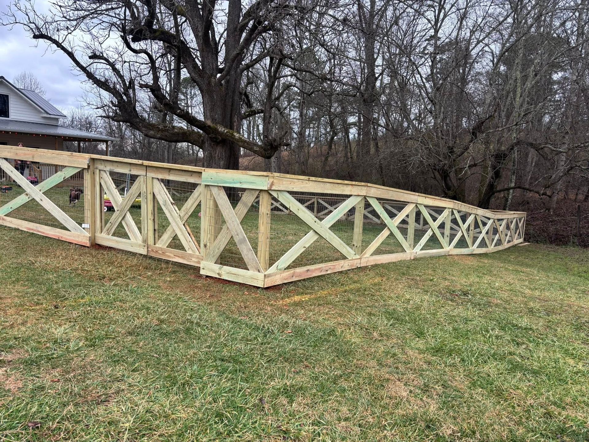 A newly constructed wooden fence with an X-pattern design stands in a grassy yard near a house and bare trees.