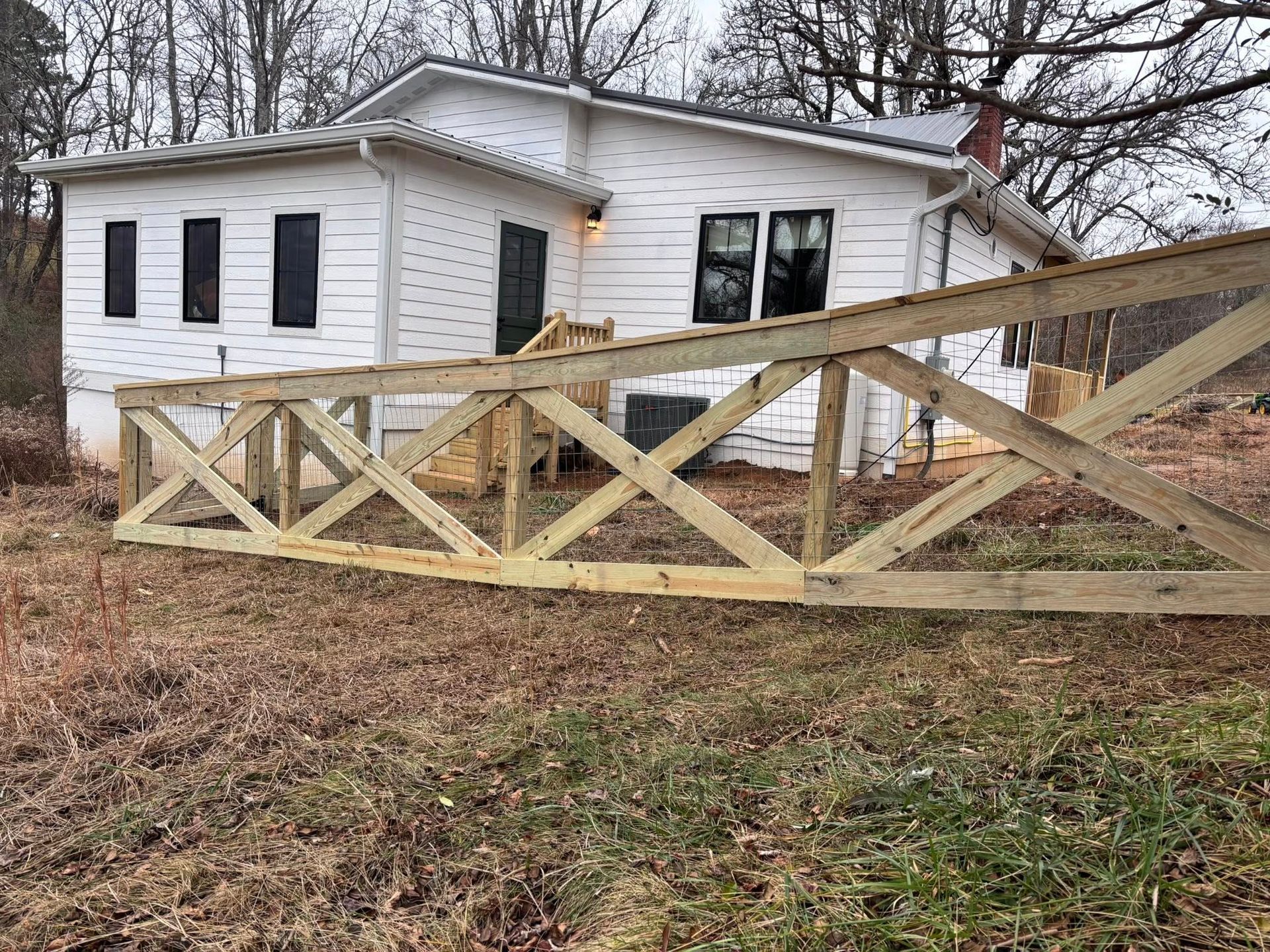 A white wooden house with a newly constructed, unfinished wooden truss fence in the foreground.