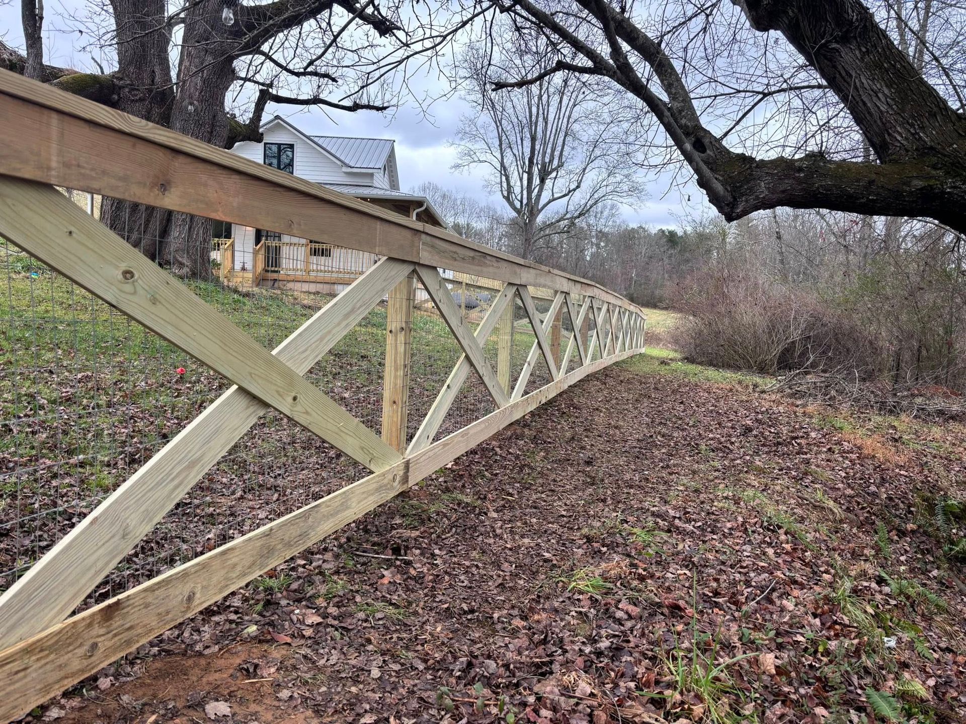 A wooden fence with X-shaped cross-bracing runs through a grassy, leaf-covered yard toward a house in the distance.