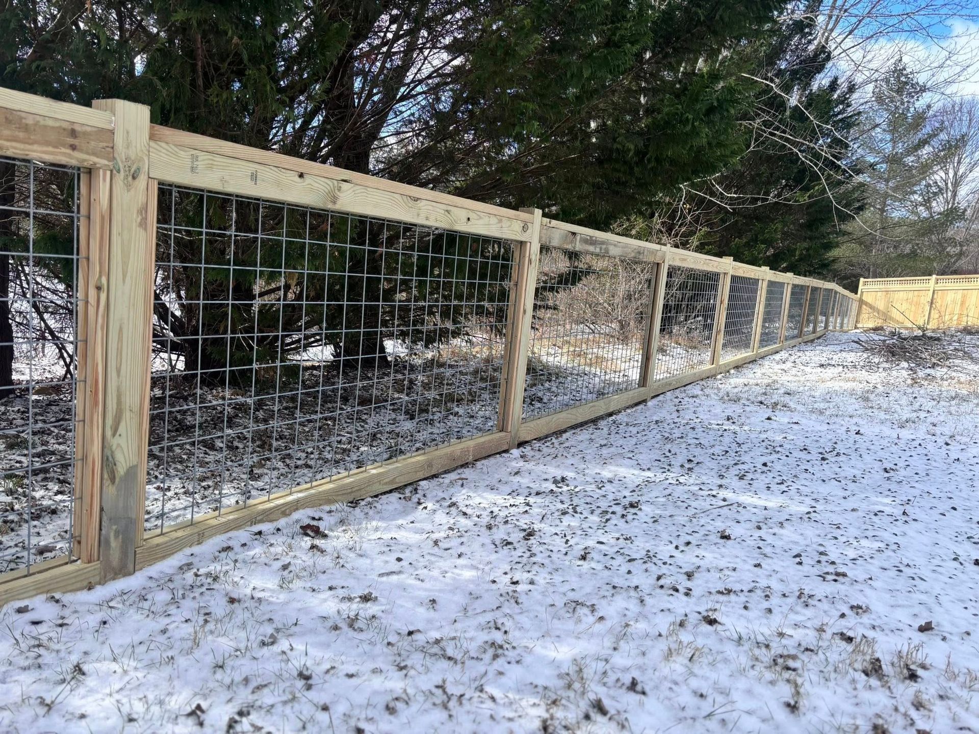 A wooden rail fence with heavy-duty wire mesh installed along a snow-covered yard next to a wooded area.