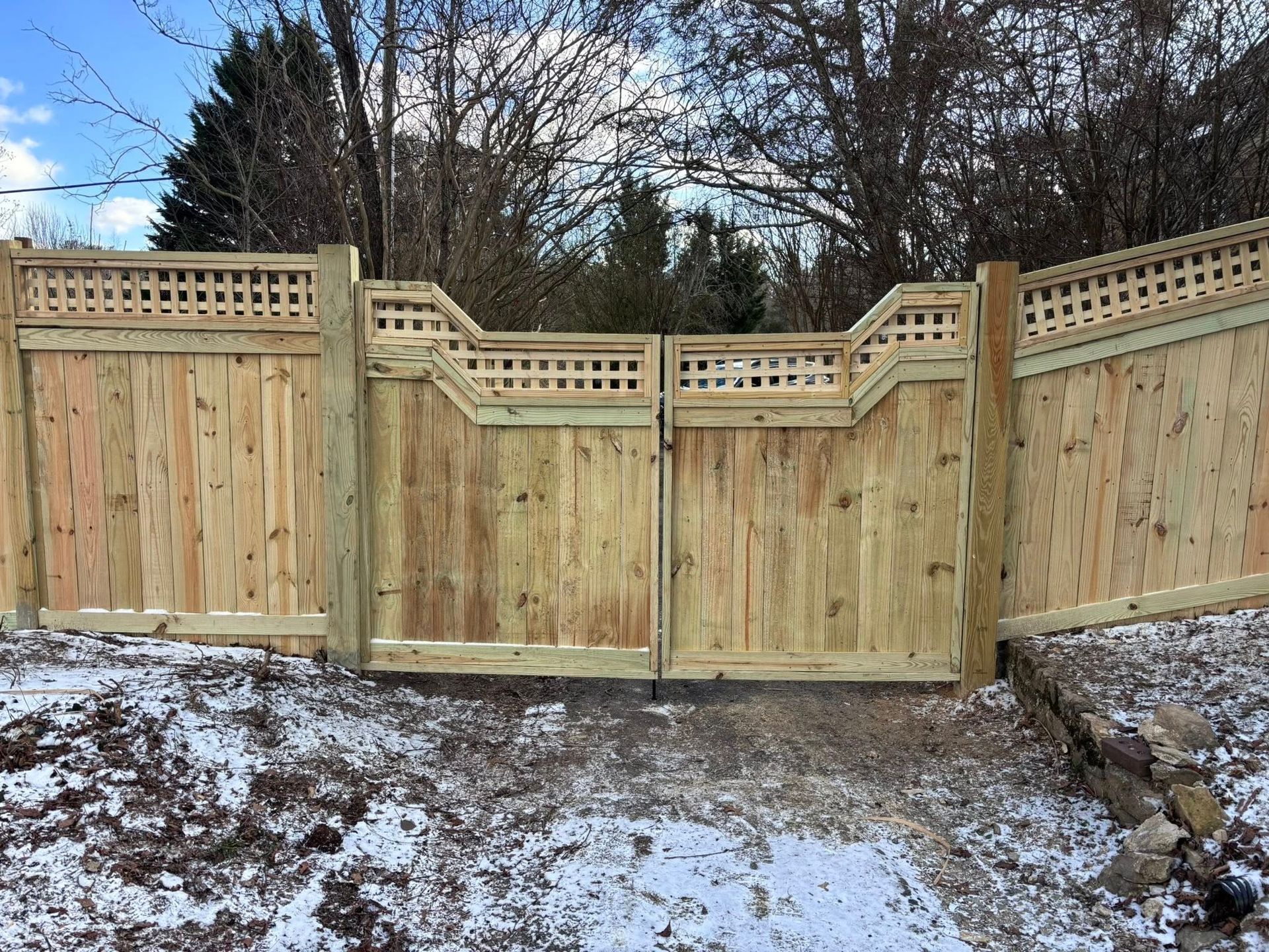 A wide wooden double gate with a decorative lattice top, centered in a matching fence line on a snowy, wooded property.