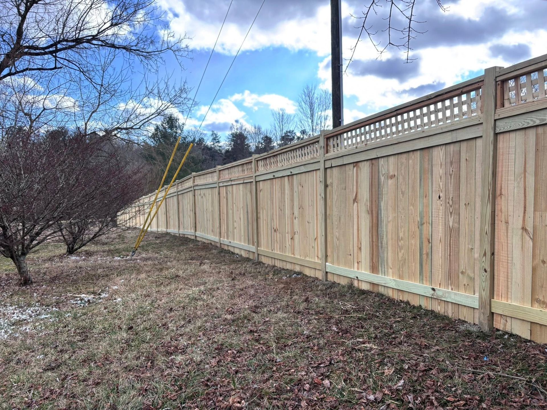 A long, wooden privacy fence with a lattice top section runs through a yard with bare trees under a cloudy sky.