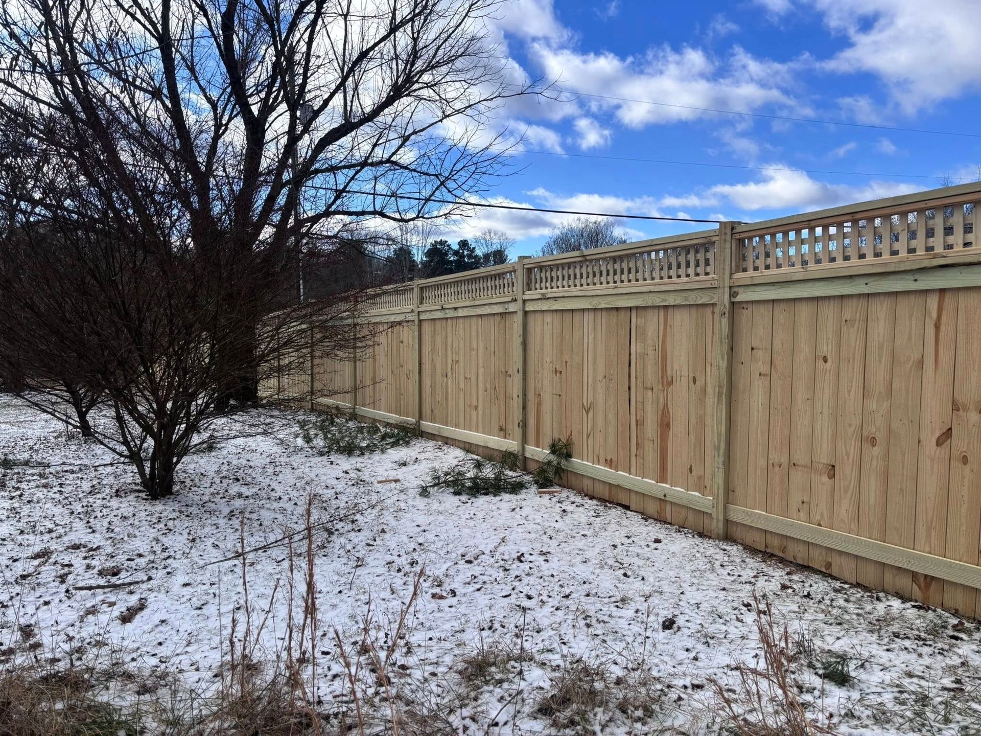 A tall wooden privacy fence with a lattice top border stands on snow-covered ground near a tree under a blue sky.