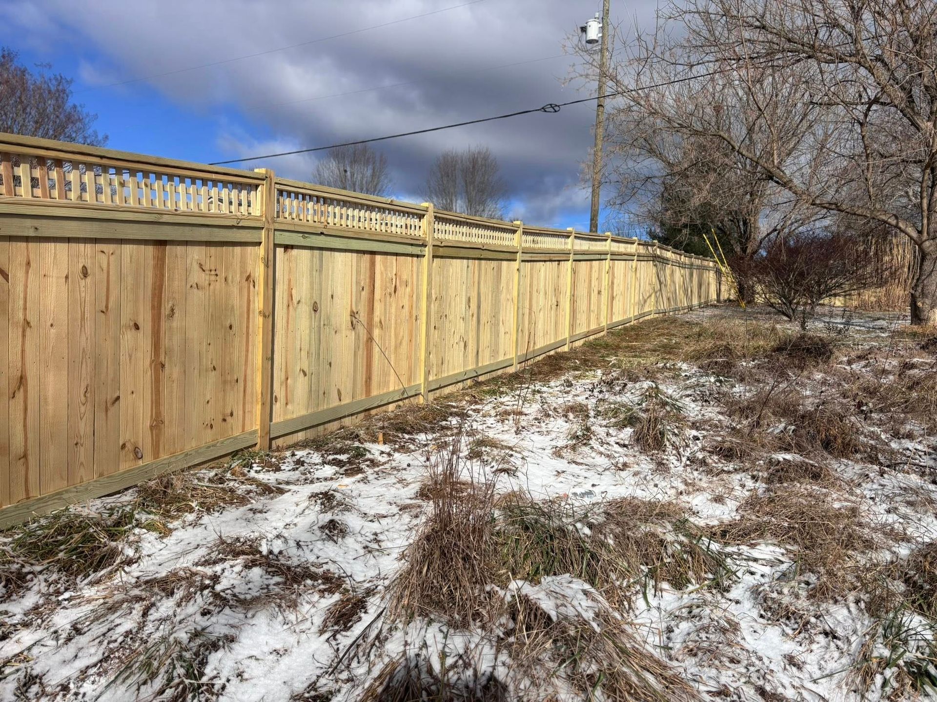 A new wooden privacy fence with decorative lattice top stretches across a snowy, unmaintained outdoor yard.