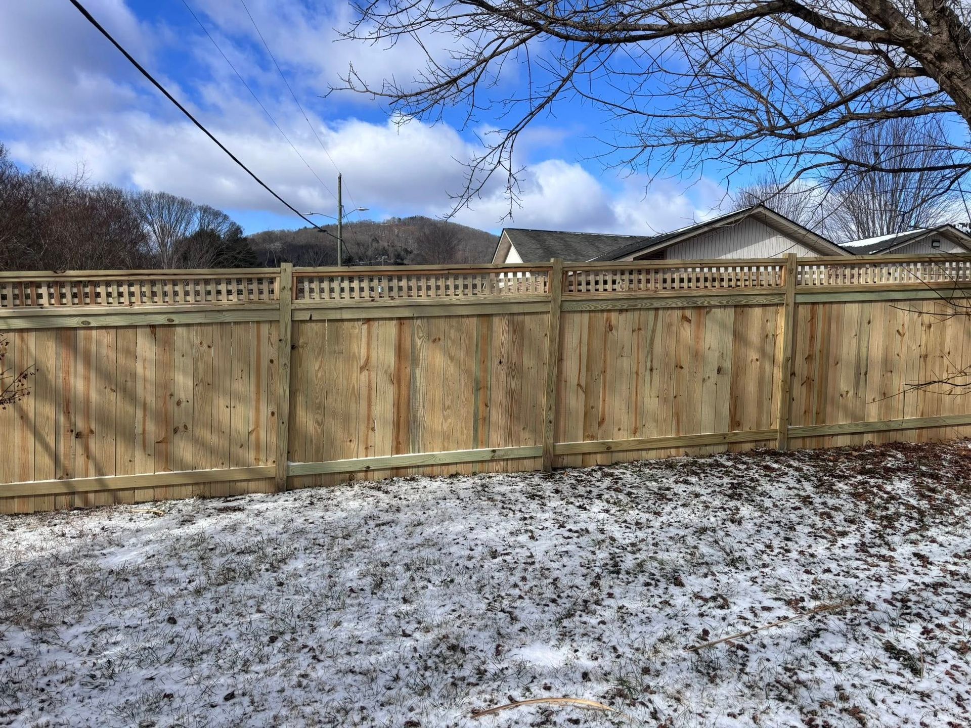 A wooden privacy fence with a lattice top border, set against a backdrop of a snowy yard, trees, and distant hills.