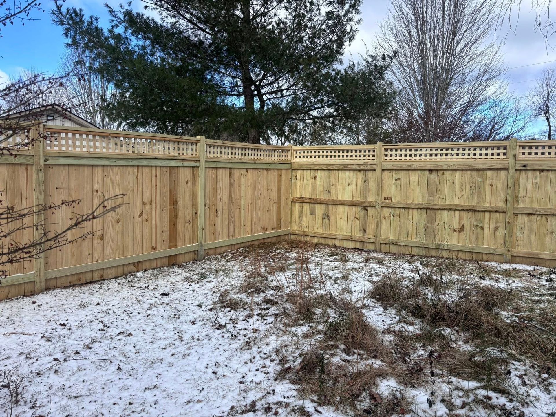 A wooden fence with a lattice top border in a backyard with patchy snow and dry vegetation.