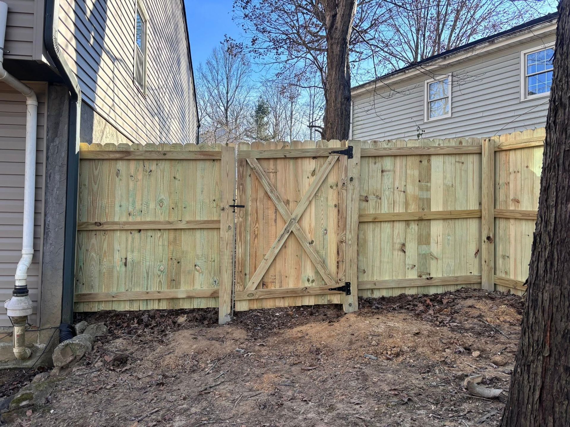 A new wooden privacy fence with an X-braced gate stands between two houses in a yard covered with brown leaves.