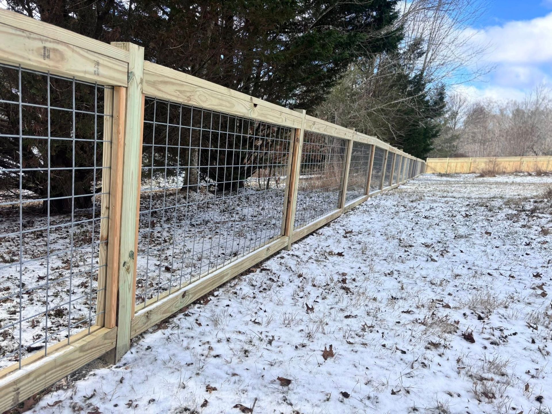 A wooden post-and-rail fence with metal grid wire panels runs through a snowy, wooded landscape.