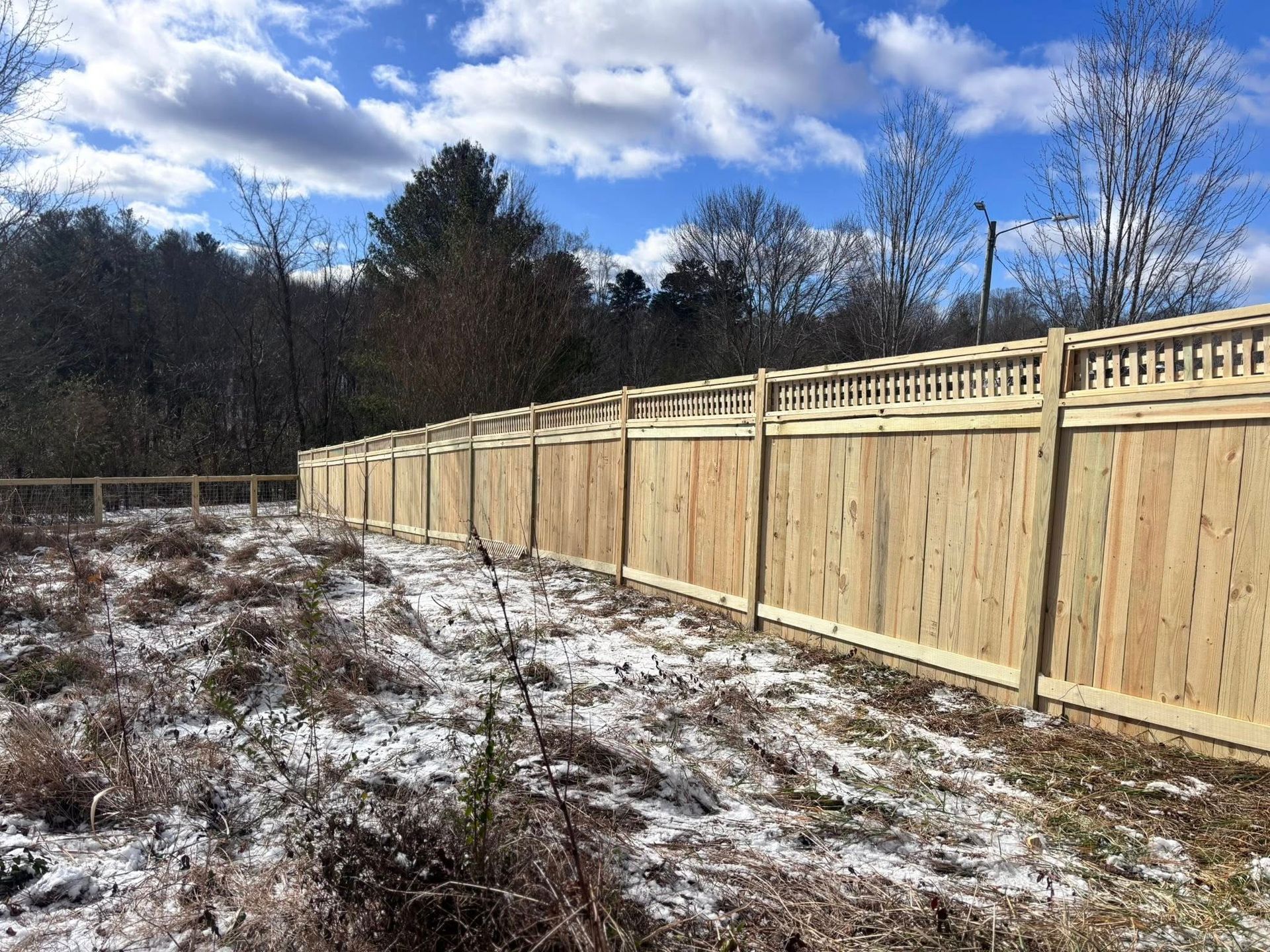 A long, wooden privacy fence with a decorative lattice top sits along a snow-dusted, grassy field under a cloudy blue sky.