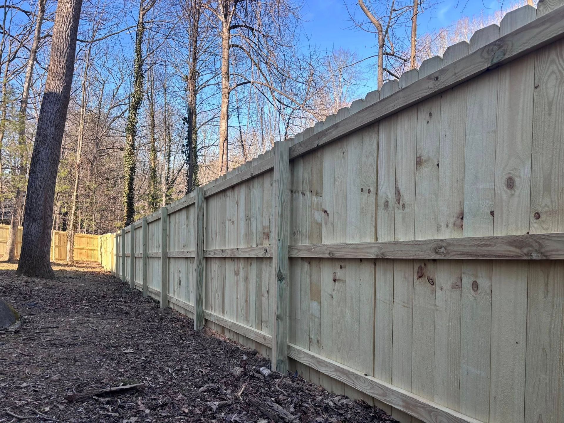 A new light-wood privacy fence runs along a wooded area covered in fallen leaves under a bright blue sky.