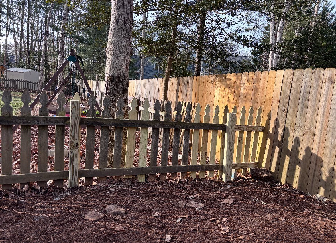 A low wooden picket fence stands in a yard, with a taller privacy fence and trees in the background.