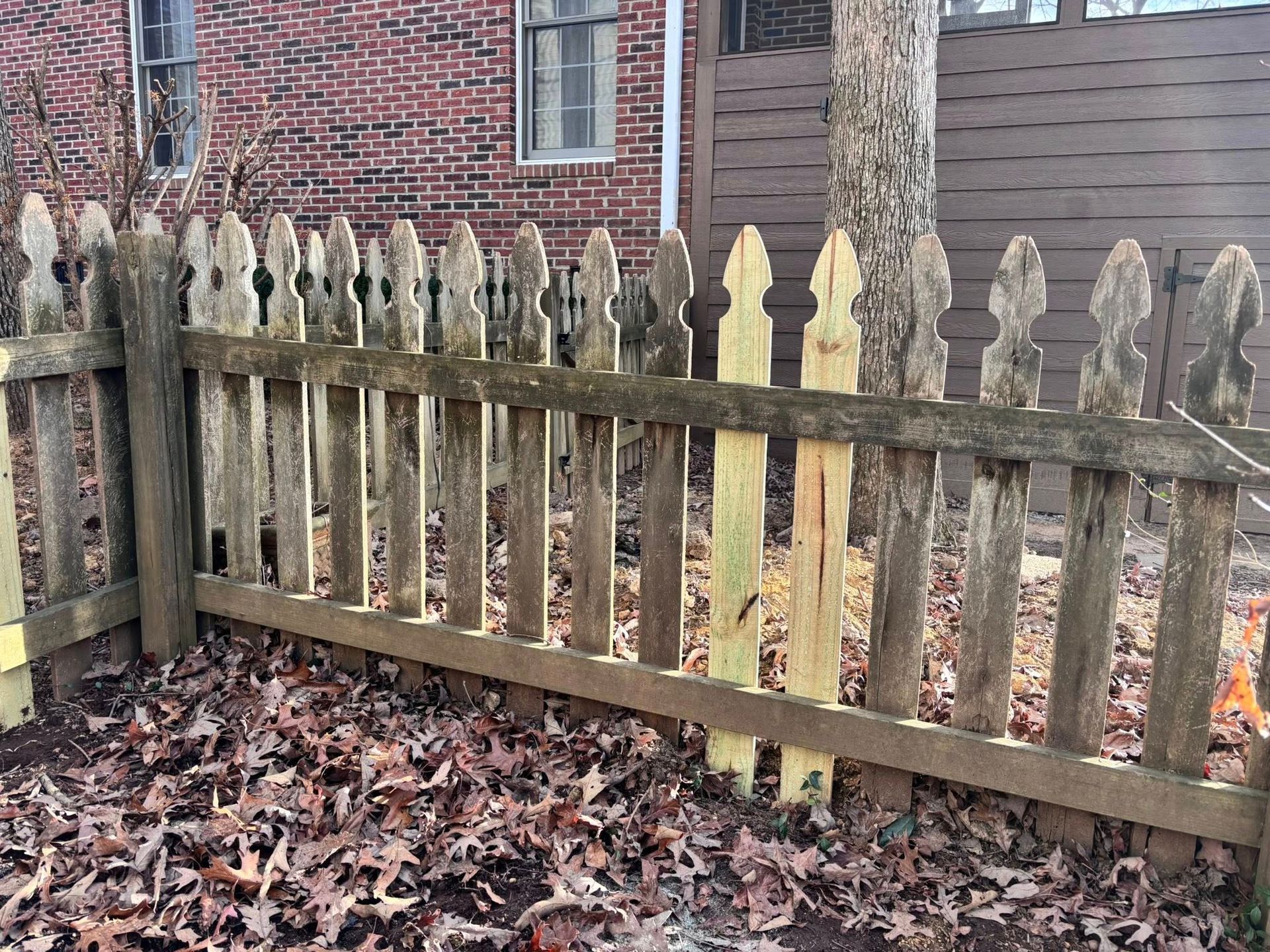 A wooden picket fence in front of a brick house, featuring two light-colored, newly replaced pickets amidst weathered ones.