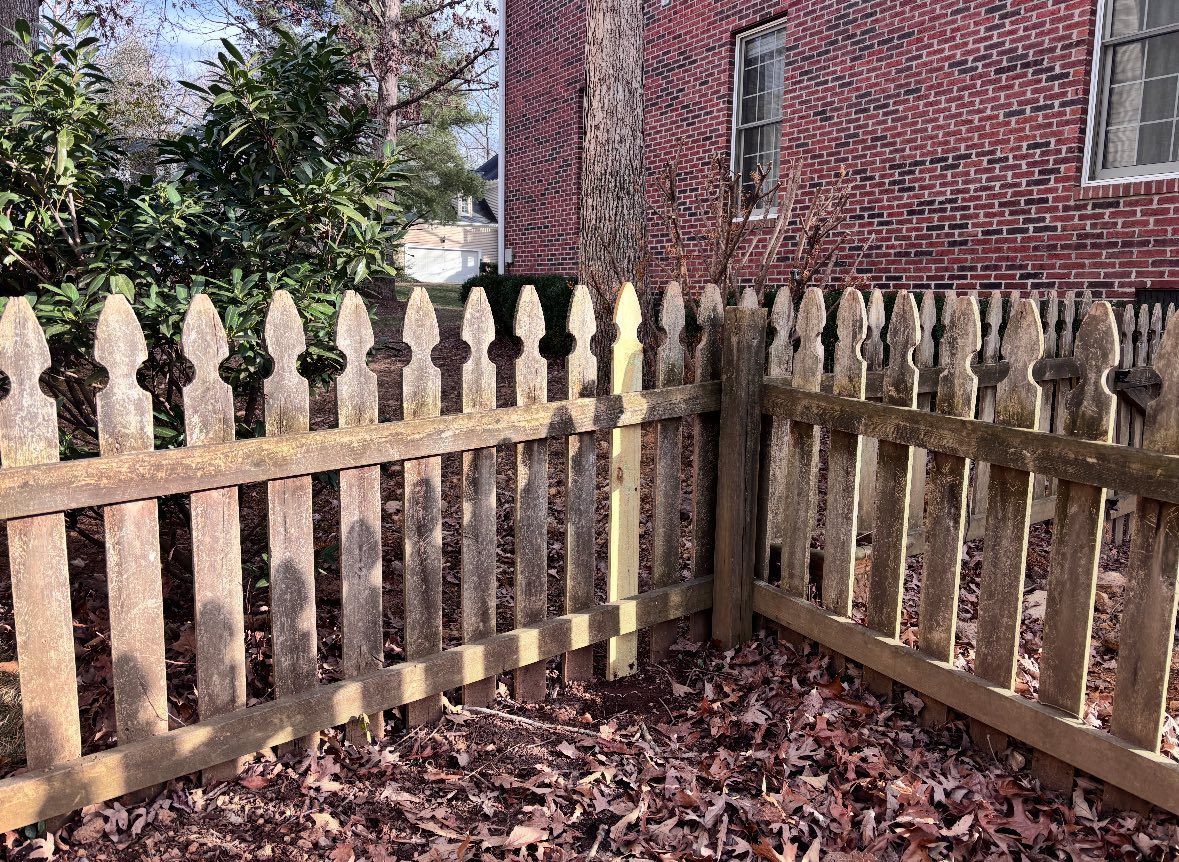 A weathered wooden picket fence in an outdoor setting, featuring a single, newer, light-colored replacement post.
