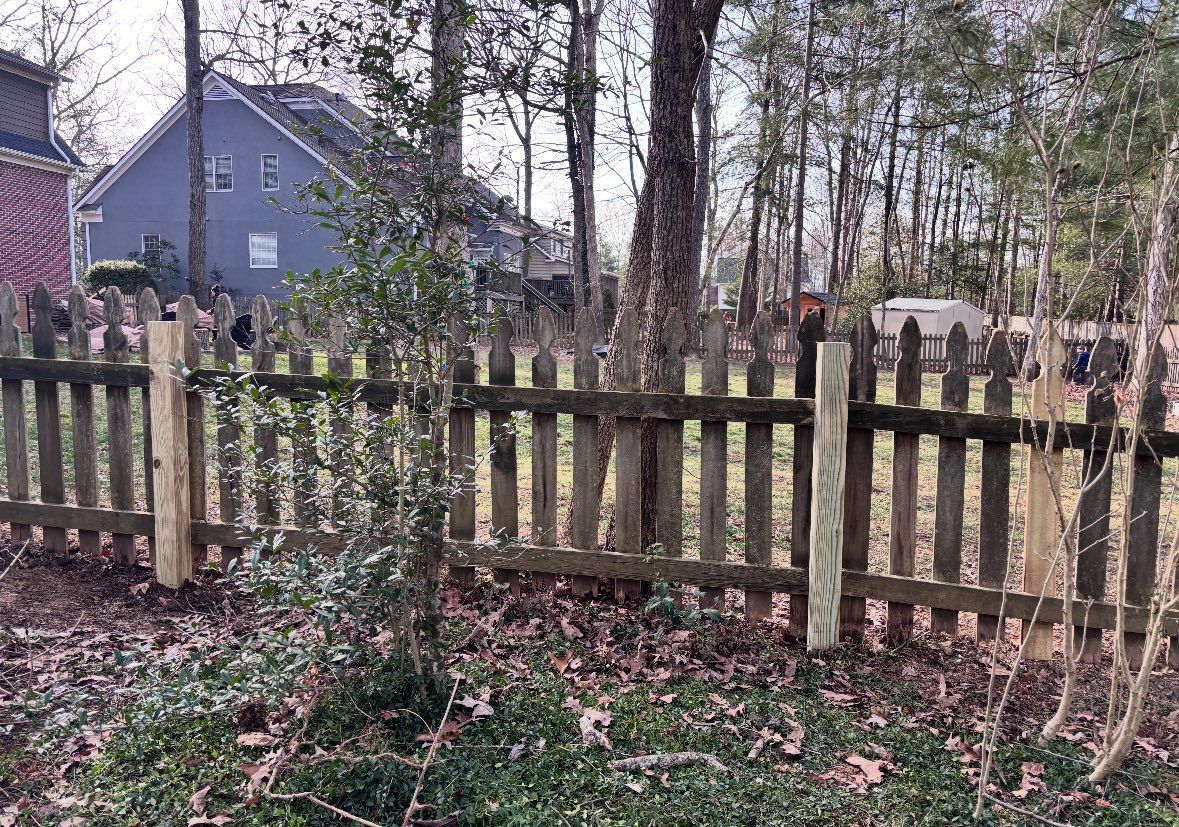 A wooden picket fence sits in a yard with scattered fallen leaves, with a blue house and trees in the background.