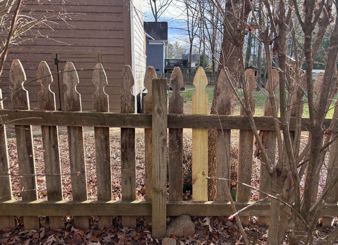 A wooden picket fence with one new, light-colored replacement picket stands among older, weathered gray fence pickets.