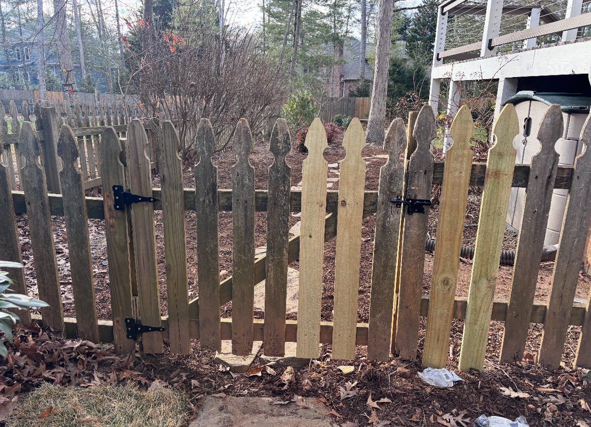 A weathered wooden picket fence with a gate, featuring two newer, light-colored replacement pickets in the middle.