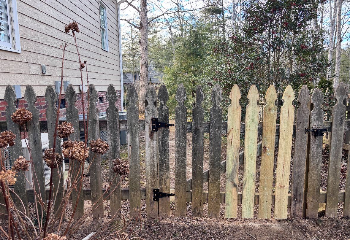 A weathered wooden picket fence in a yard features two light-colored, recently replaced pickets amidst the aged wood.