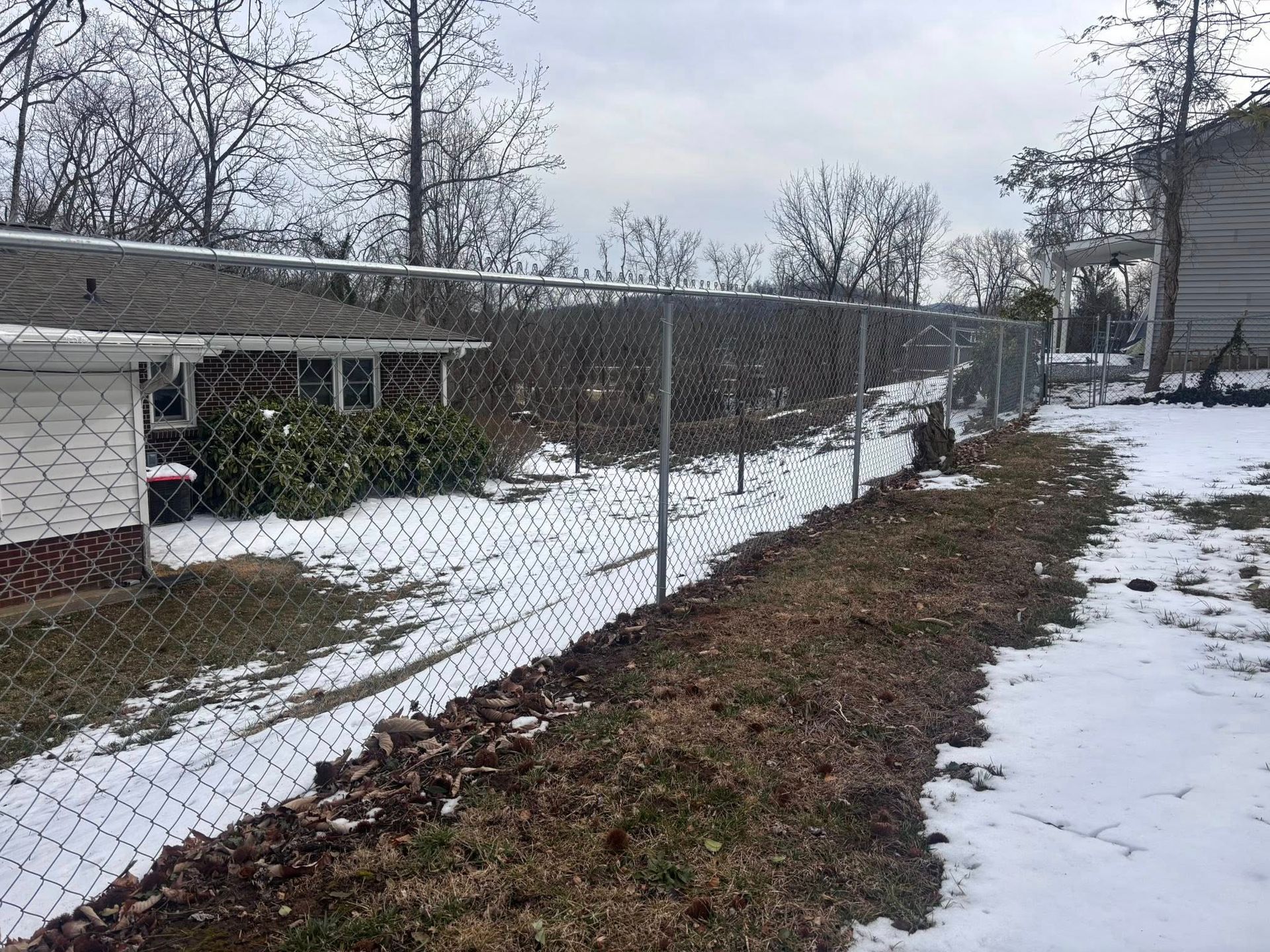 A chain-link fence divides a snow-covered yard from a nearby house on a cloudy day.