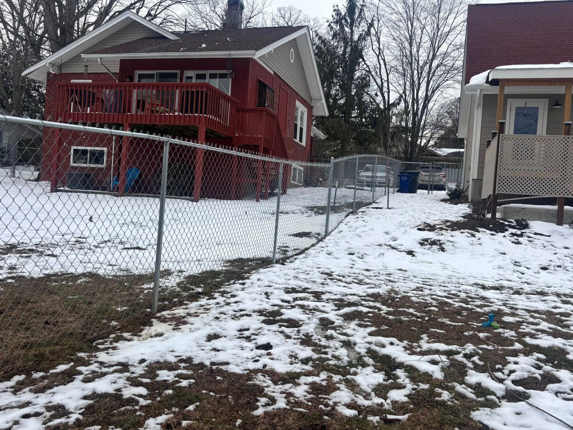 A chain-link fence divides a snow-covered backyard between a red house with a deck and a neighboring grey house.