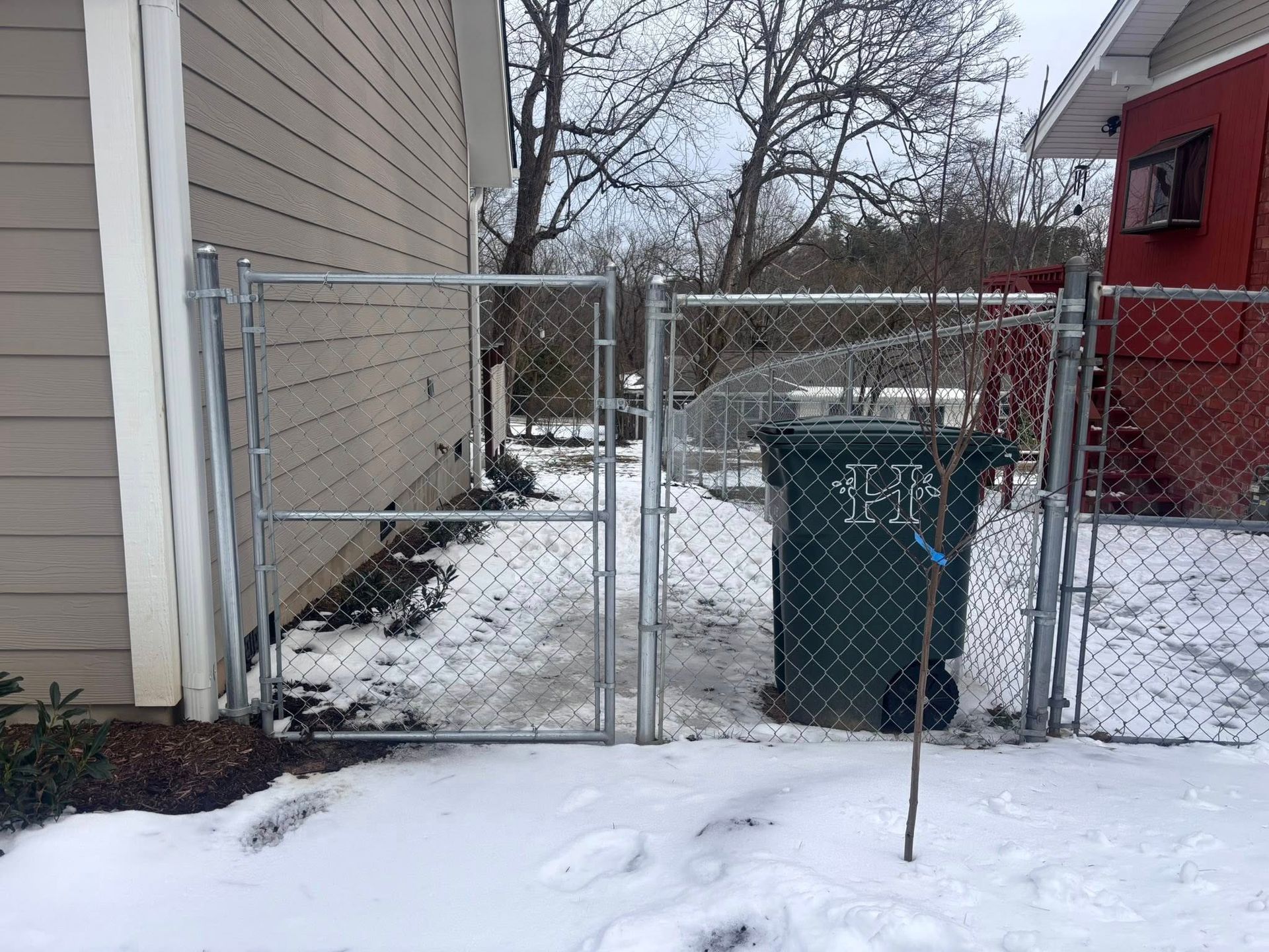 A chain-link fence and gate between a tan house and a red building, with a green trash bin on snow-covered ground.