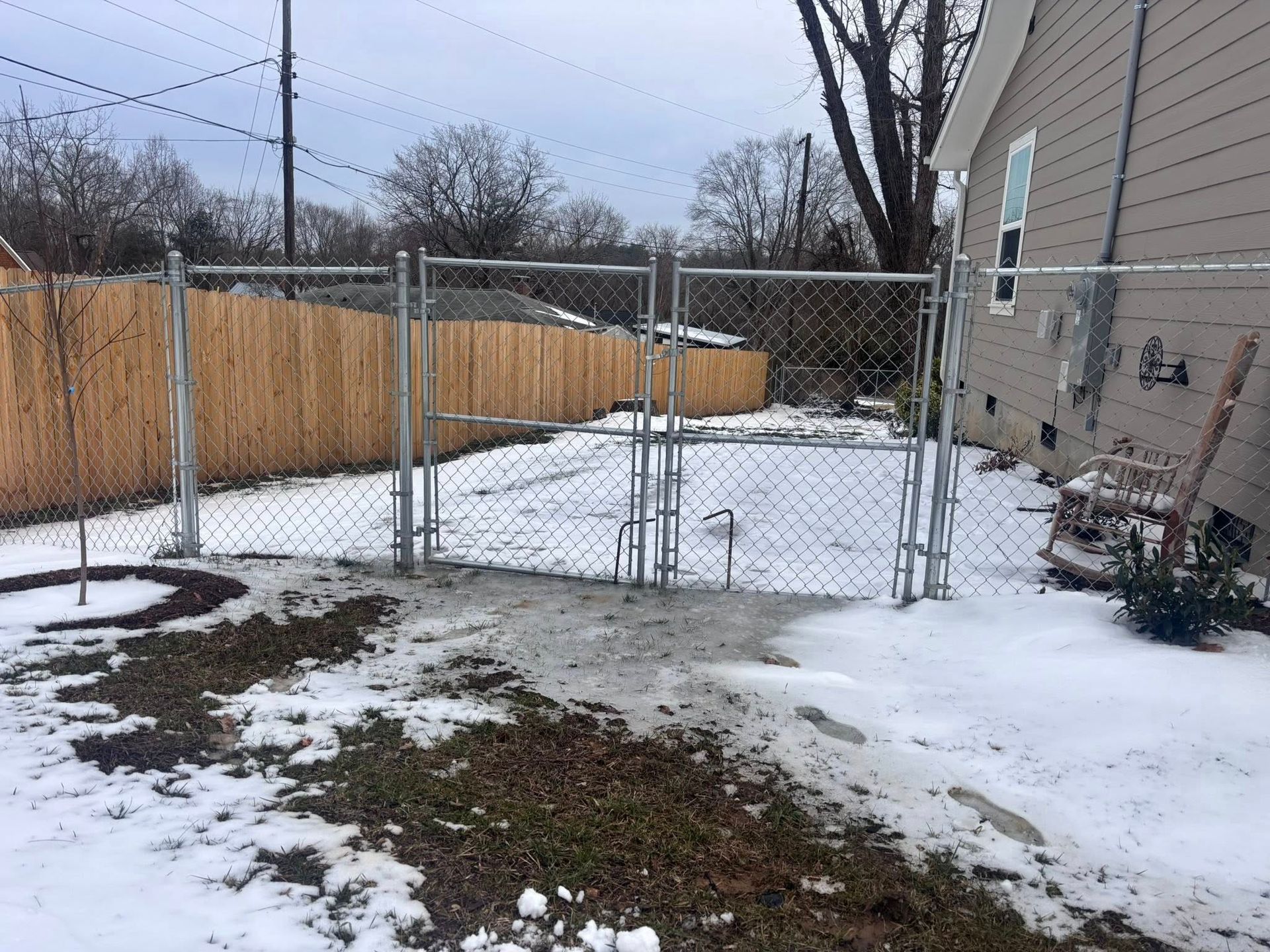 A chain-link fence gate closes off a snow-covered backyard next to a residential house with beige siding.