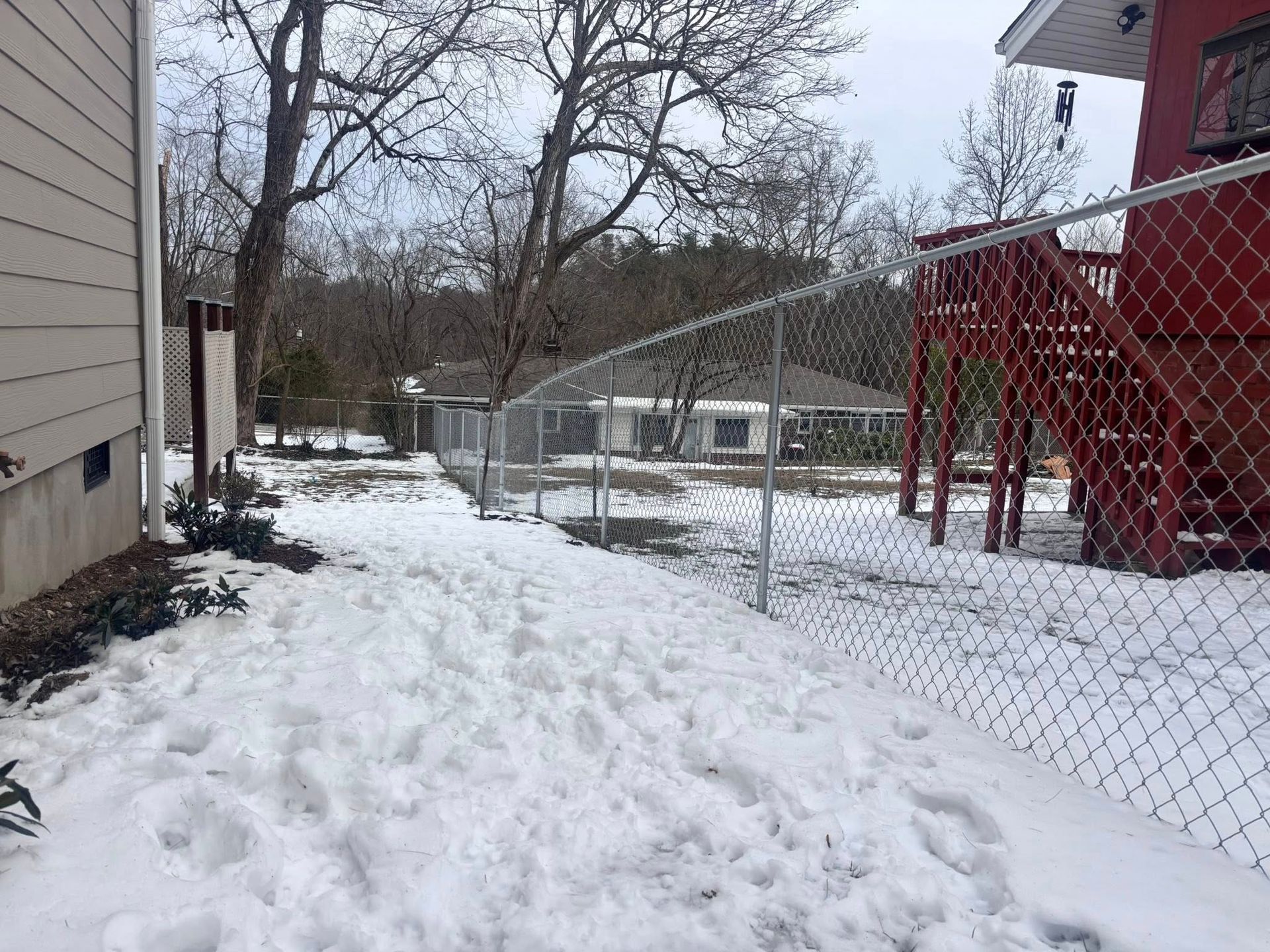 A snowy backyard features a chain-link fence separating a grassy area from a house with a red deck and staircase.