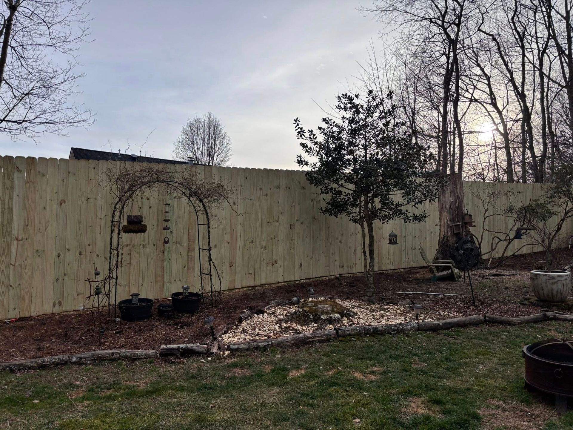 A wooden privacy fence borders a backyard with an arched garden trellis, a small tree, and a mulch garden bed.