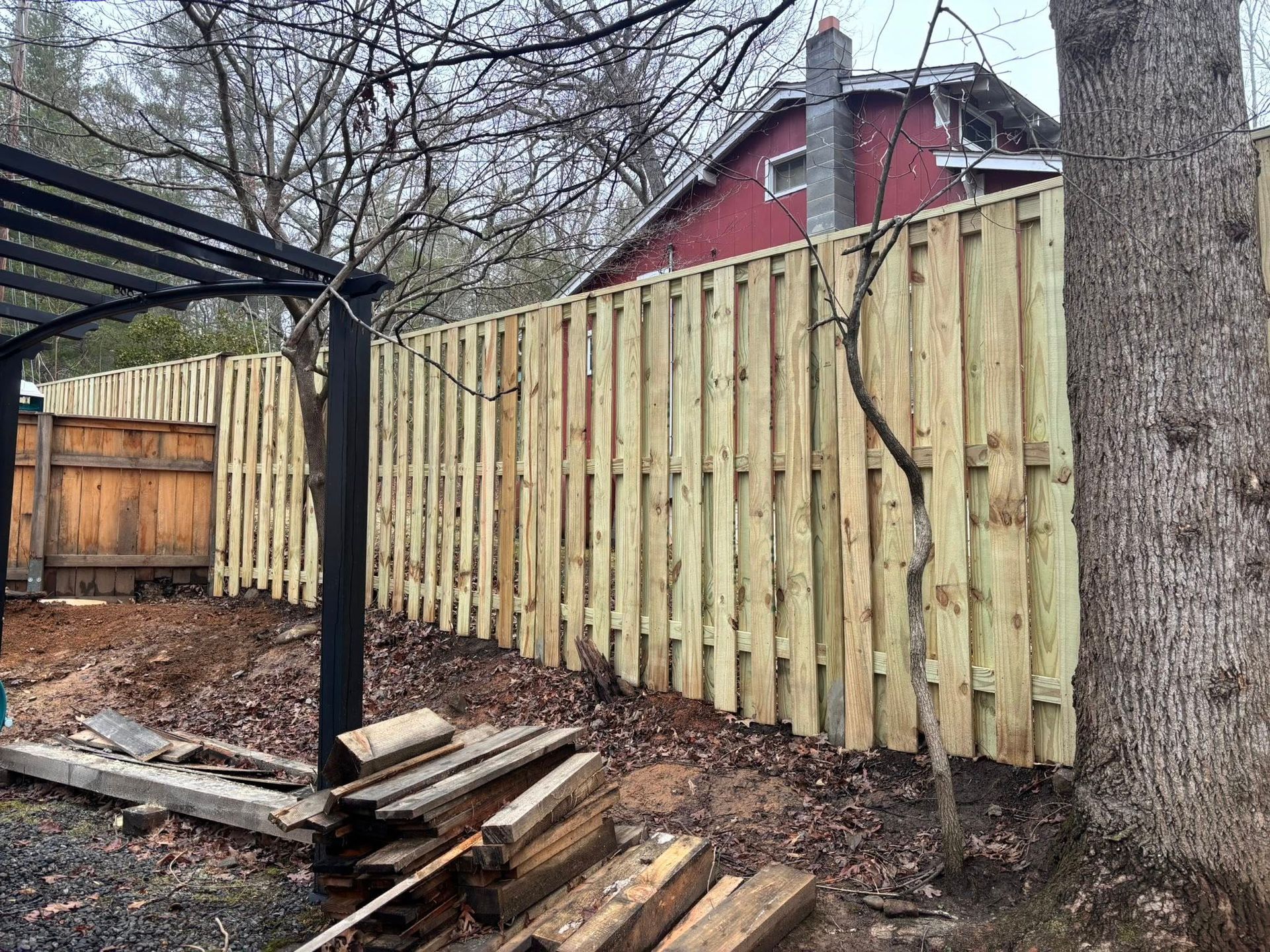 A new light-colored wood privacy fence spans a yard near a pergola and a red house under a cloudy sky.