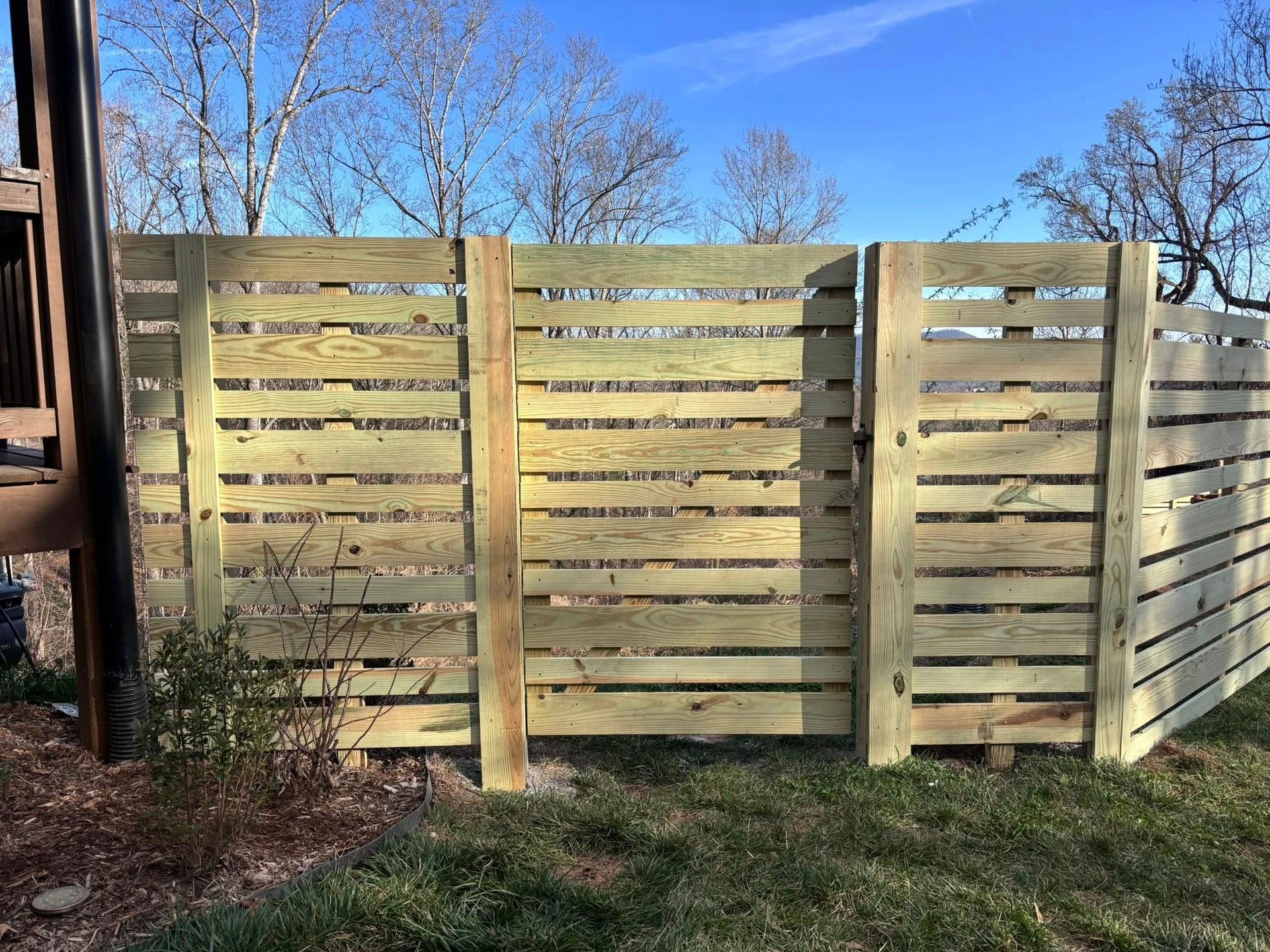 A horizontal wood slat privacy fence stands in a grassy yard under a clear blue sky.
