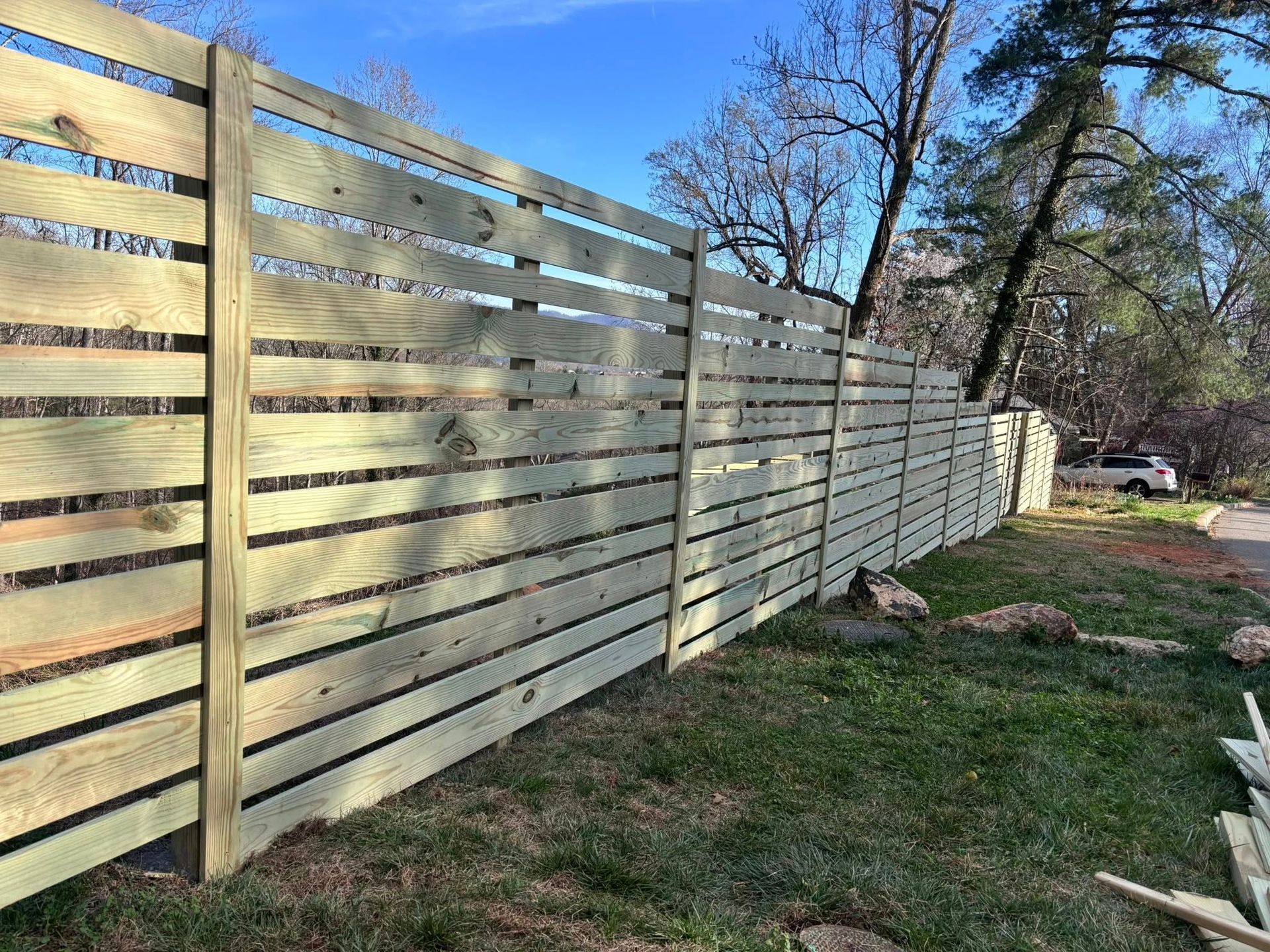 A horizontal, wooden slat fence made of light-colored, pressure-treated lumber stands in a grassy area under a blue sky.
