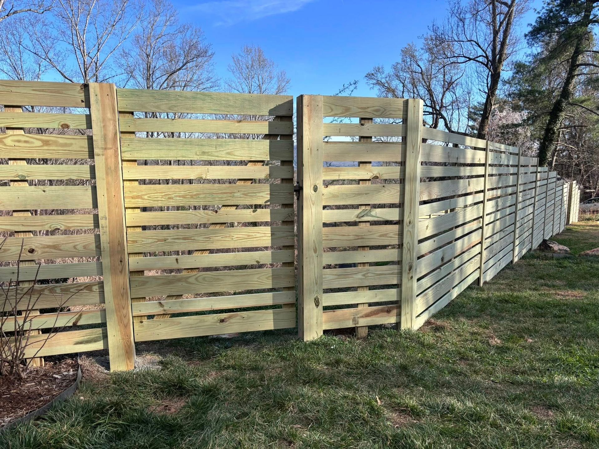 A horizontal wooden slat fence built with light-colored timber runs along a grassy lawn under a clear blue sky.