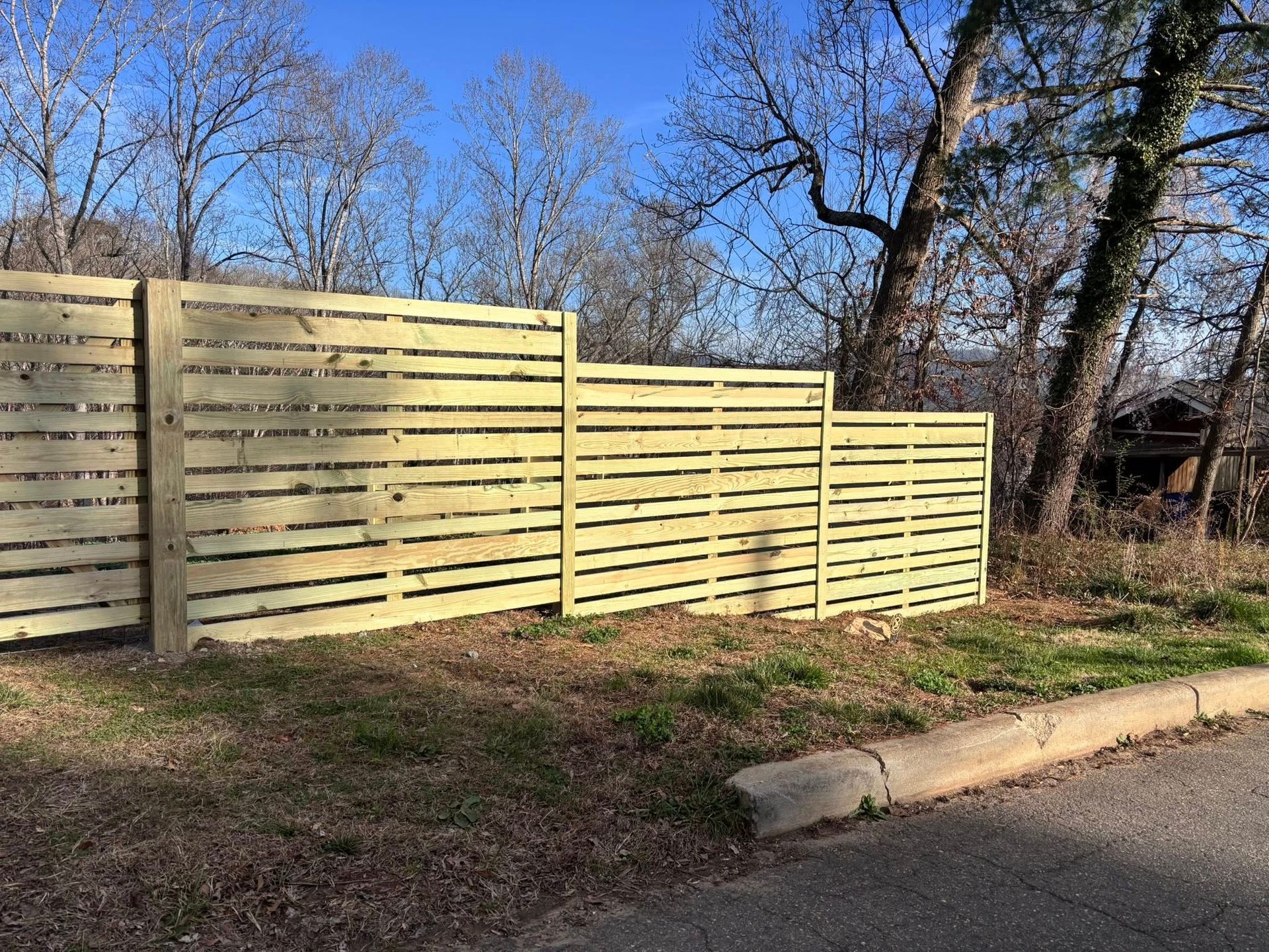 A tiered, light-colored horizontal wood fence stands in a grassy area with trees in the background under a blue sky.