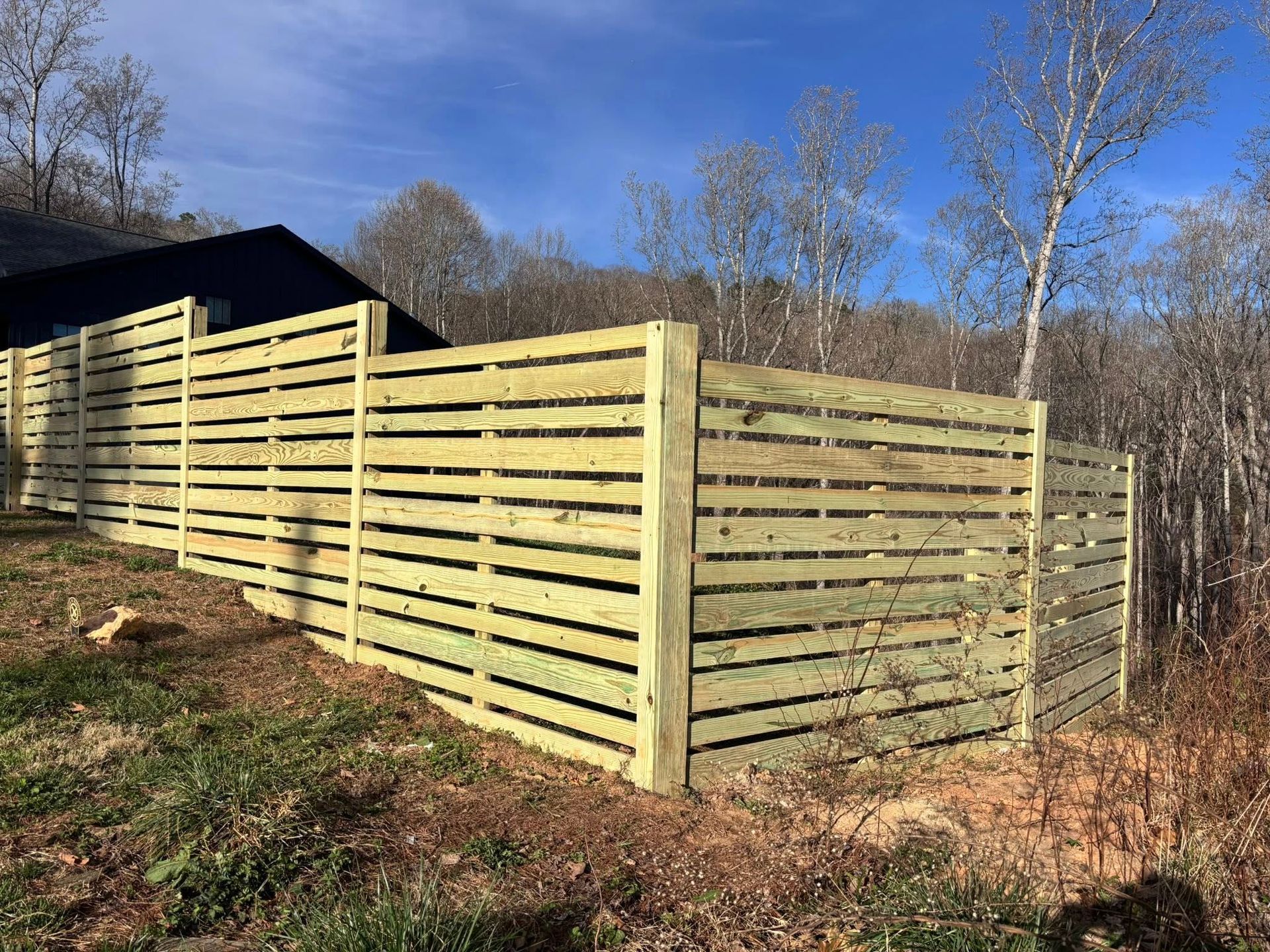 A long, light-colored horizontal wood-slat privacy fence built on a grassy hillside near a house and forest.