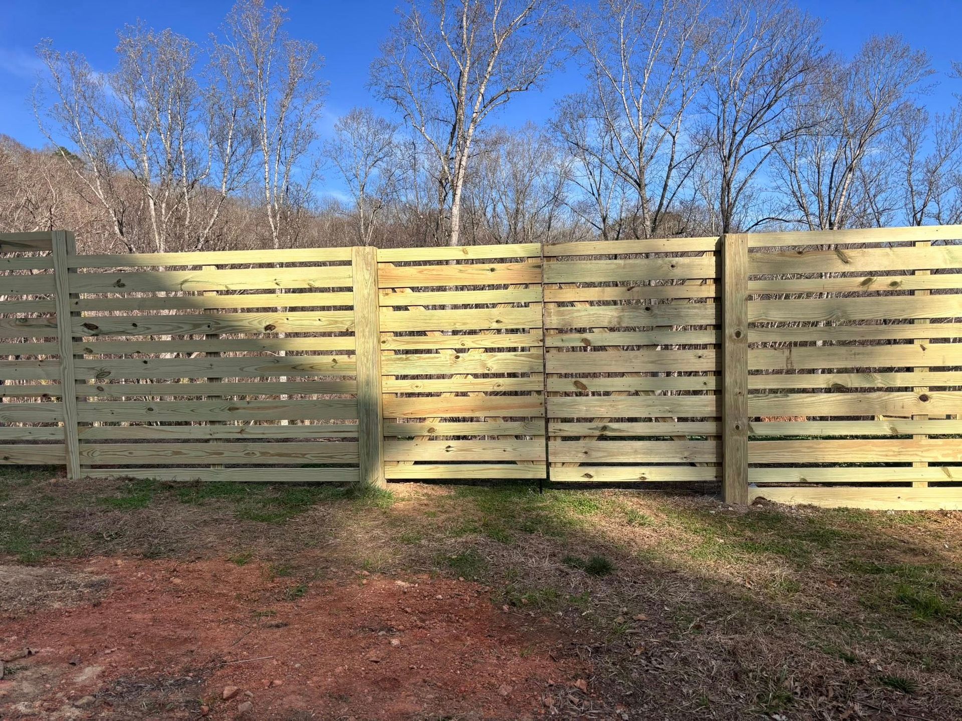 A horizontal wooden slat fence stands in a grassy, partially shaded area with bare trees against a blue sky background.